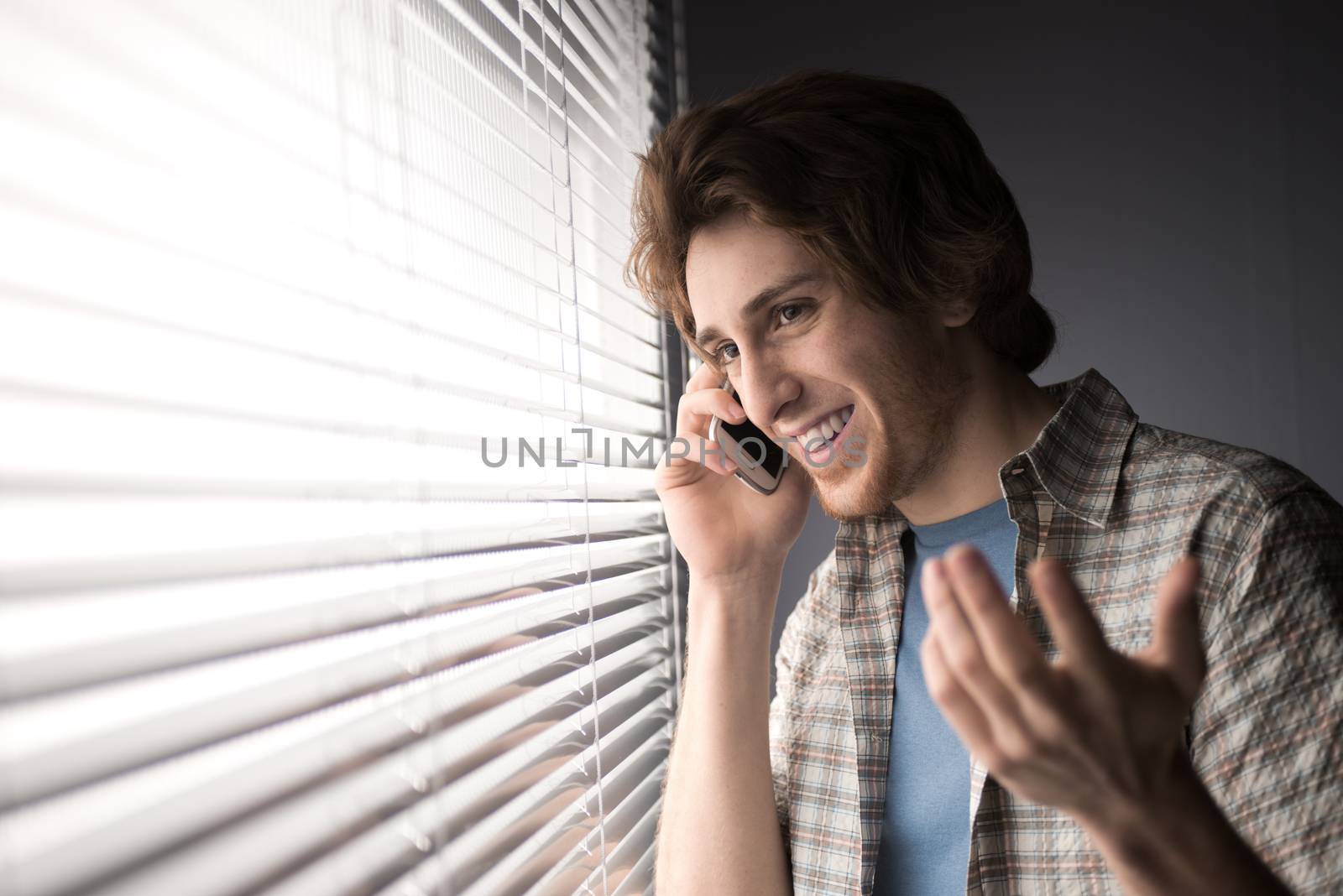 Young man smiling and talking on the phone in front of a window.