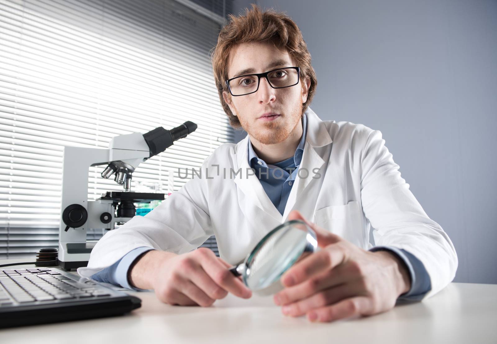 Young researcher holding a magnifier with microscope on background.