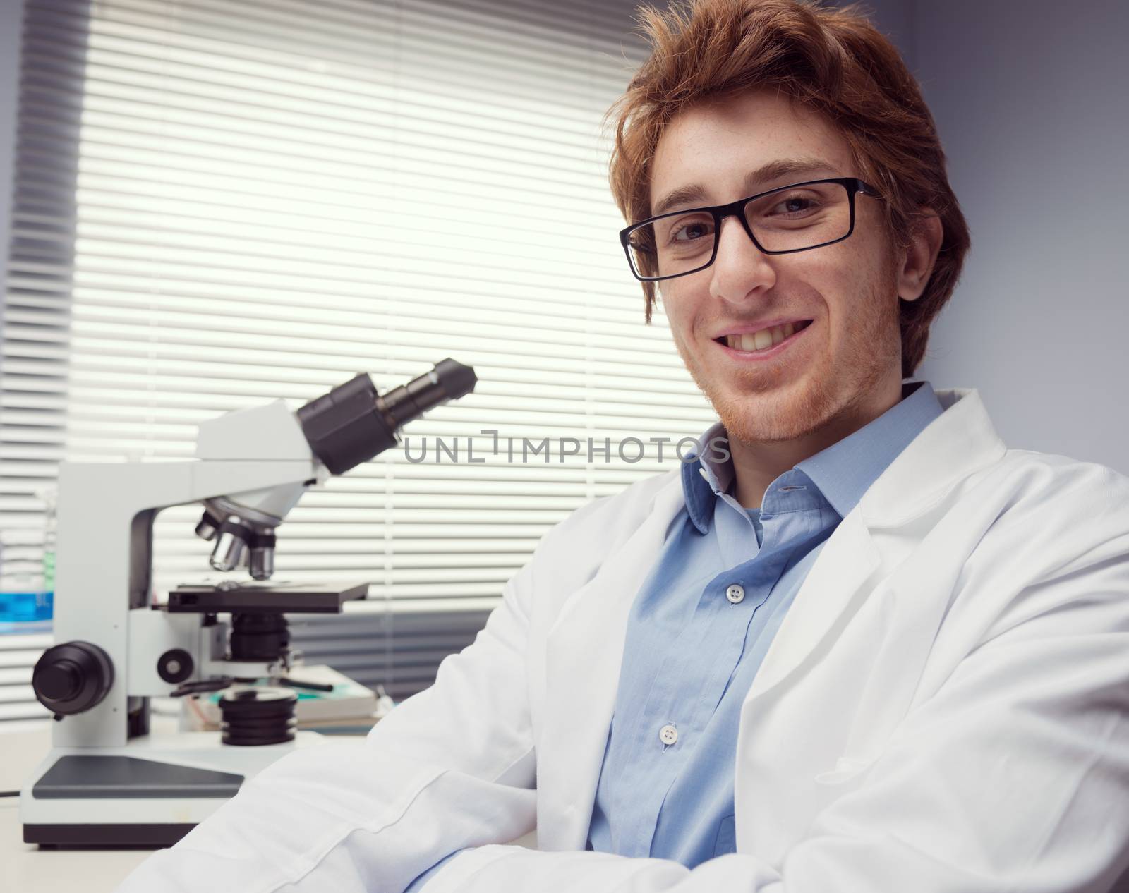 Student researcher smiling with arms crossed and microscope on background.