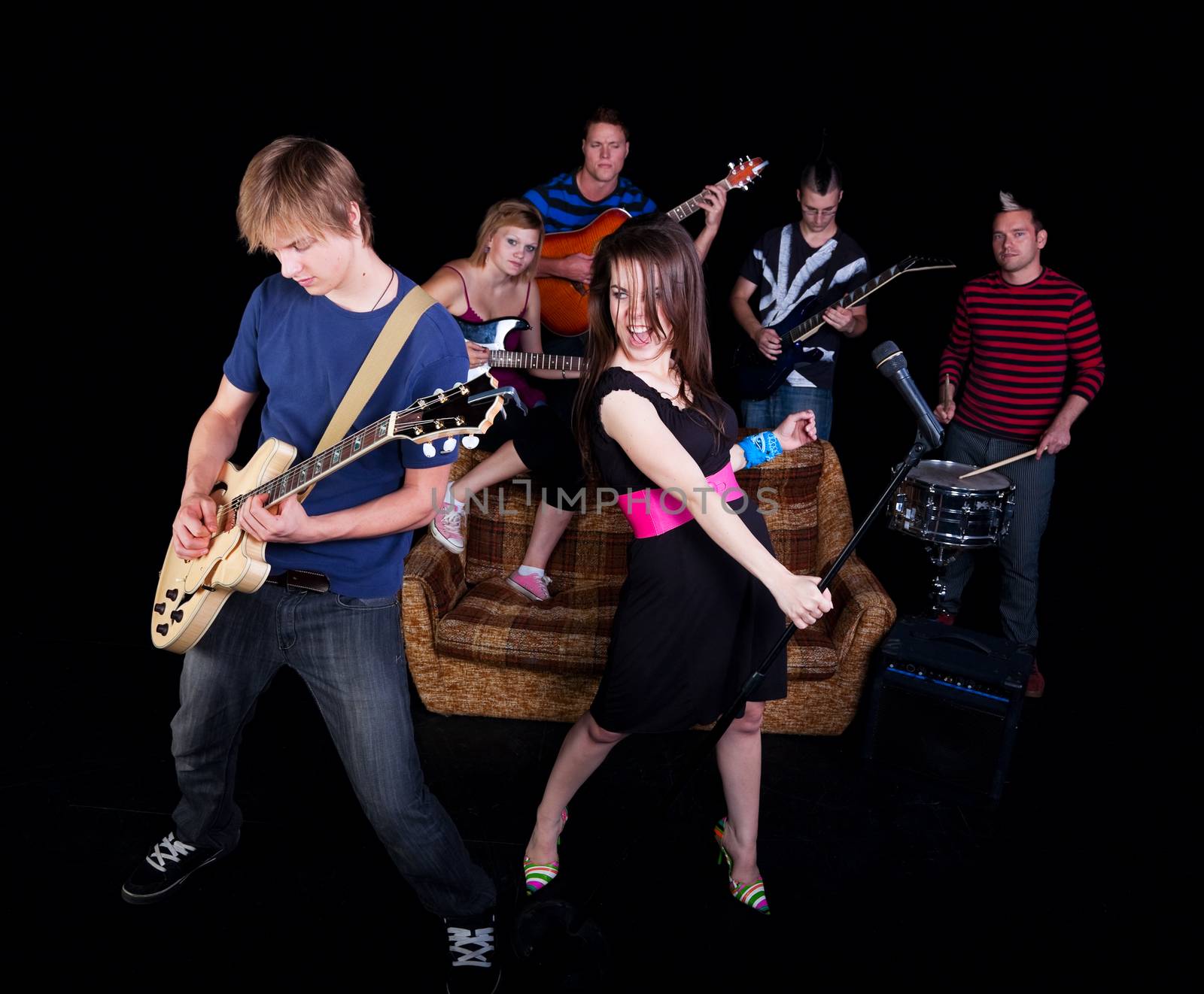 Six teens in a high school rock band practicing on a stage.