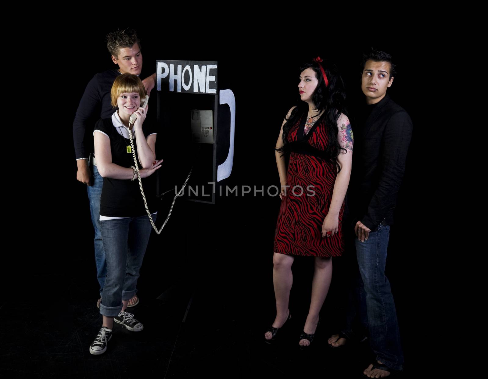 Four teenagers in a high school play.  A girl & her boyfrind wait for their turn to use a public phone while another girl chats away.