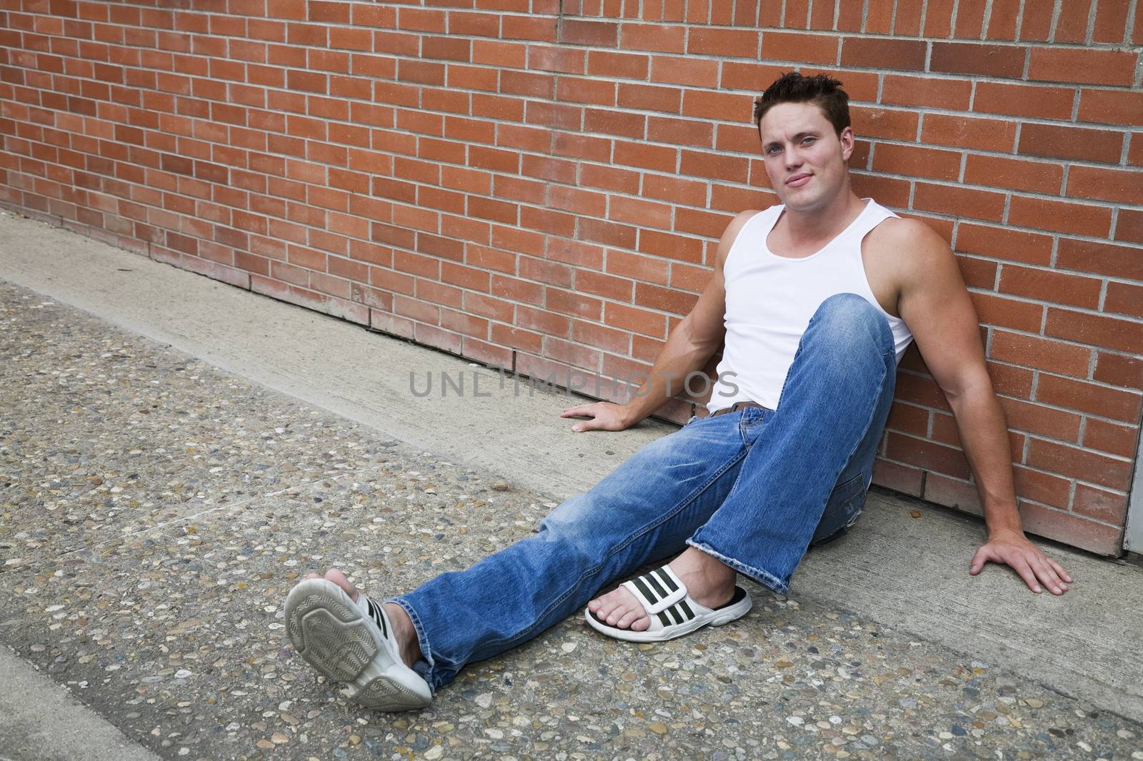 A young man sitting on a sidewalk , leaning against a brick wall.