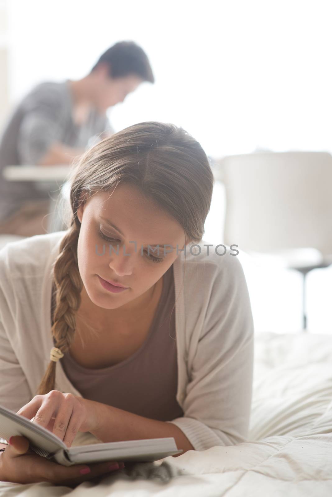 Beautiful girl reading a book while lying on bed. Boy studying on background