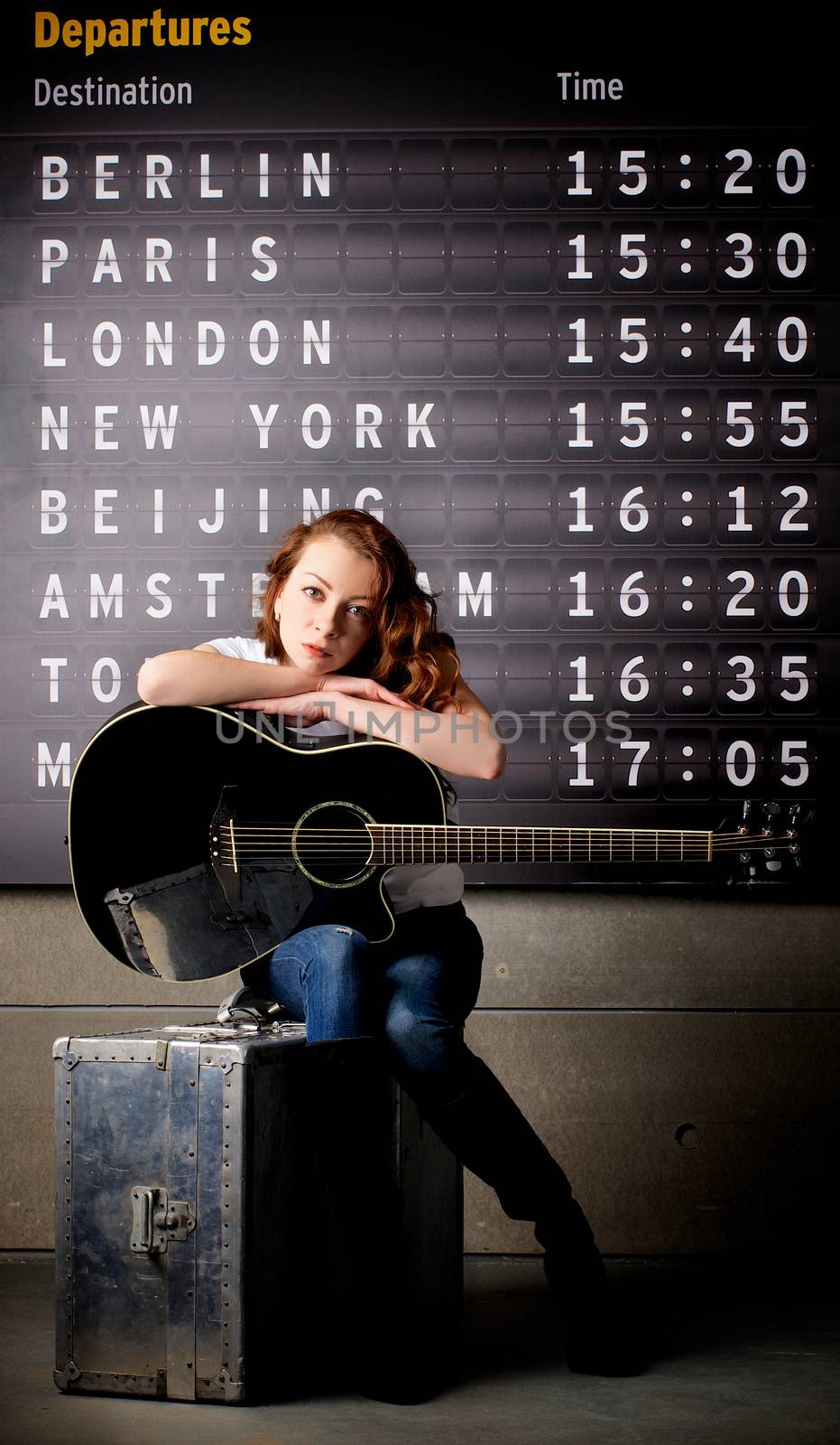 Attractive Young Woman with Guitar Sitting on Obsolete Suitcase against Arrival  Departure Board