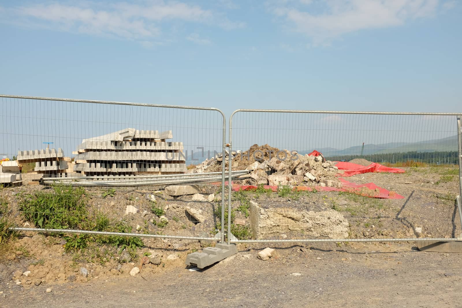 A metal fence separates development land with rubble and a stack of concrete curbstones against a blue sky with cloud