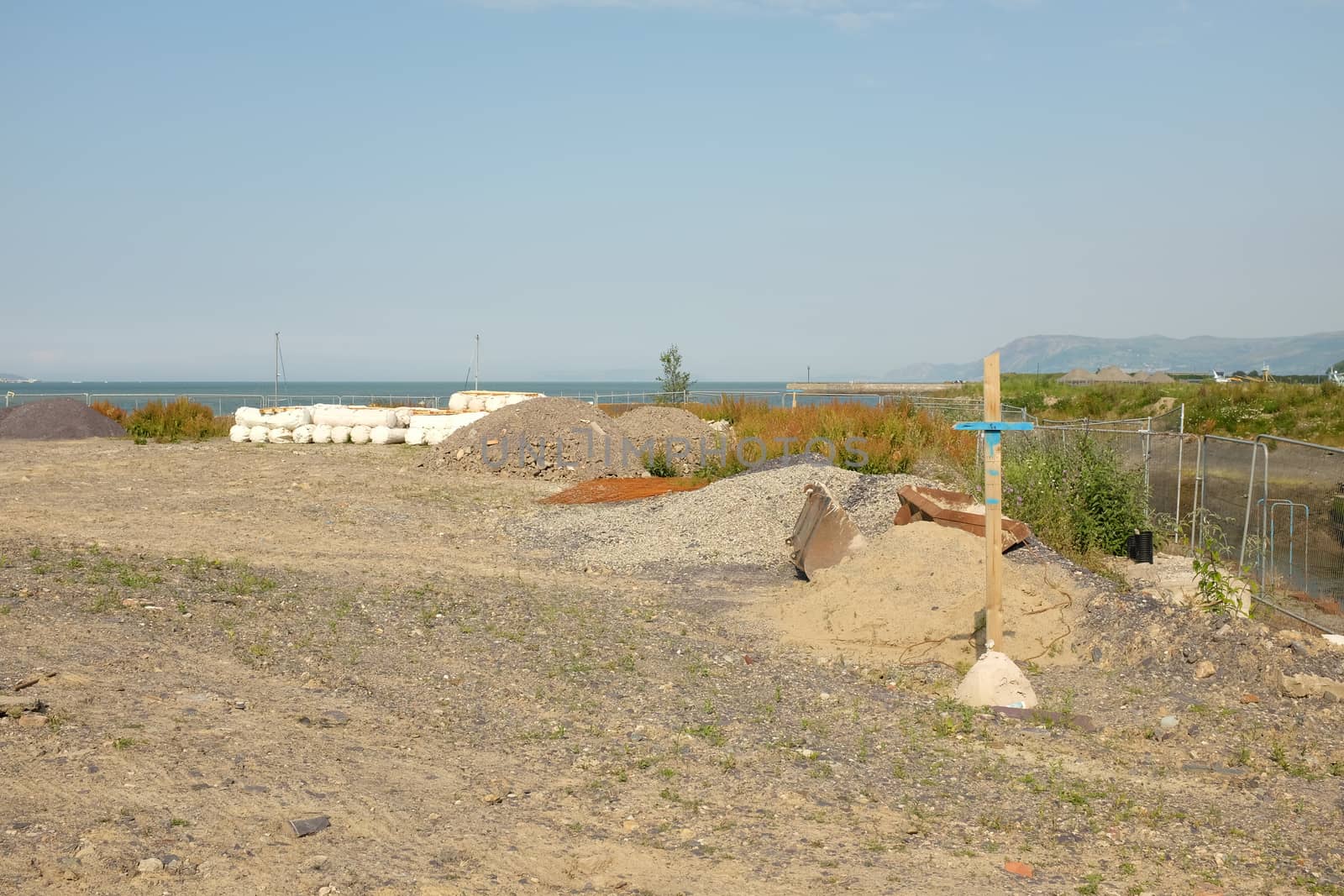 An area of cleared development land next to the sea with aggrogates and a wooden marker post.