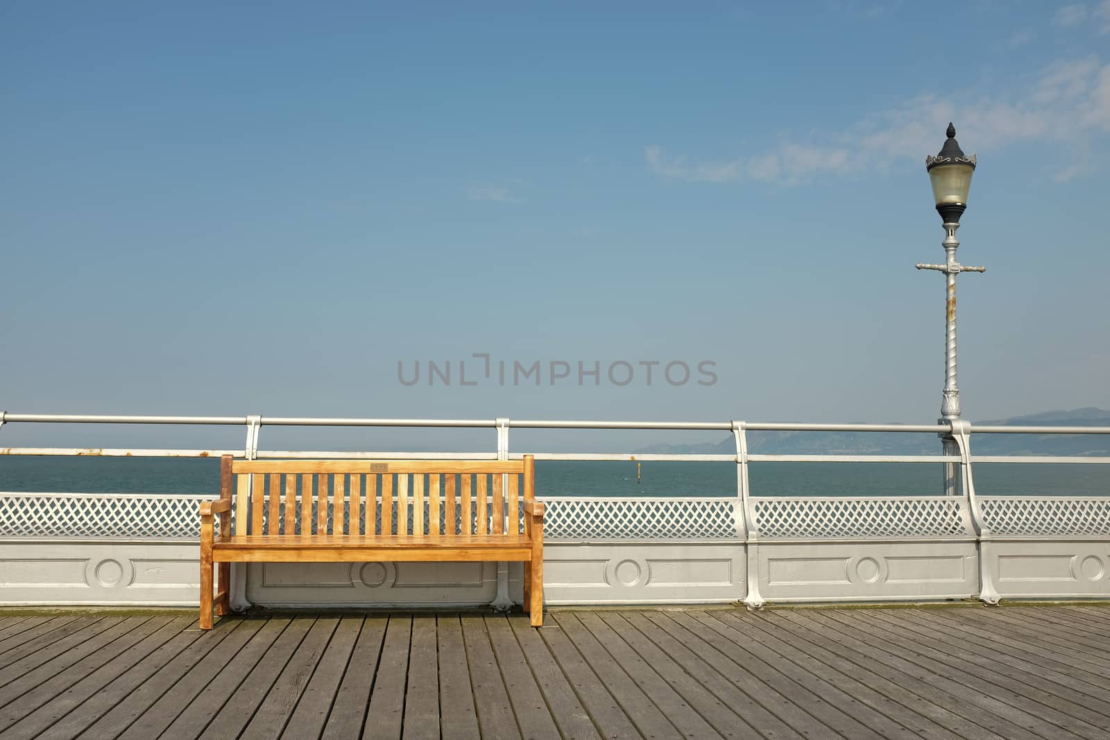 A wooden seat on a boardwalk against a silver painted railing with a light on a post against the sea and sky.