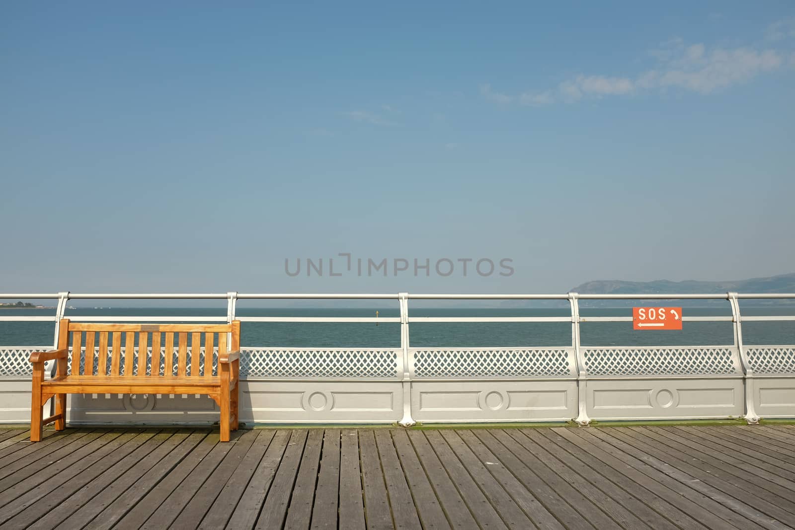 A wooden seat on a boardwalk against a silver painted railing with an orange sign with the letters S.O.S with the sea and sky in the distance.