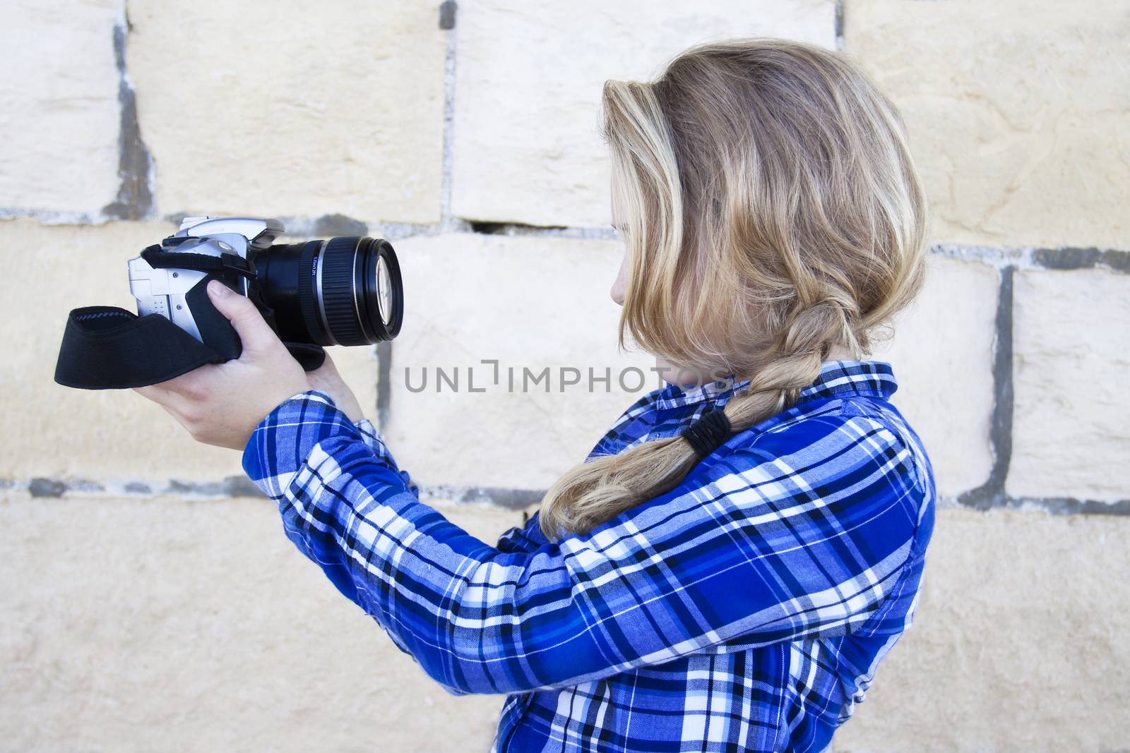 Young teenager holding up a digital SLR camera, snapping a shot of herself, in front of an old rubble wall outside