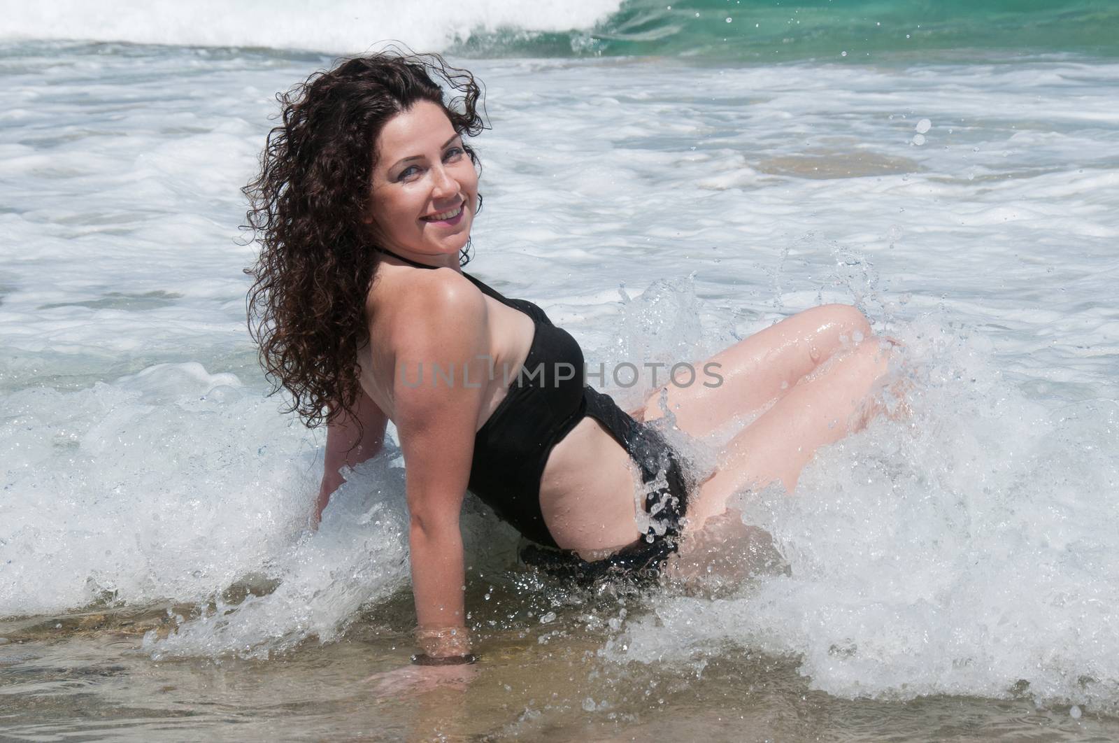 Girl in a black bathing suit sitting in the waves in the sea and laughing happily