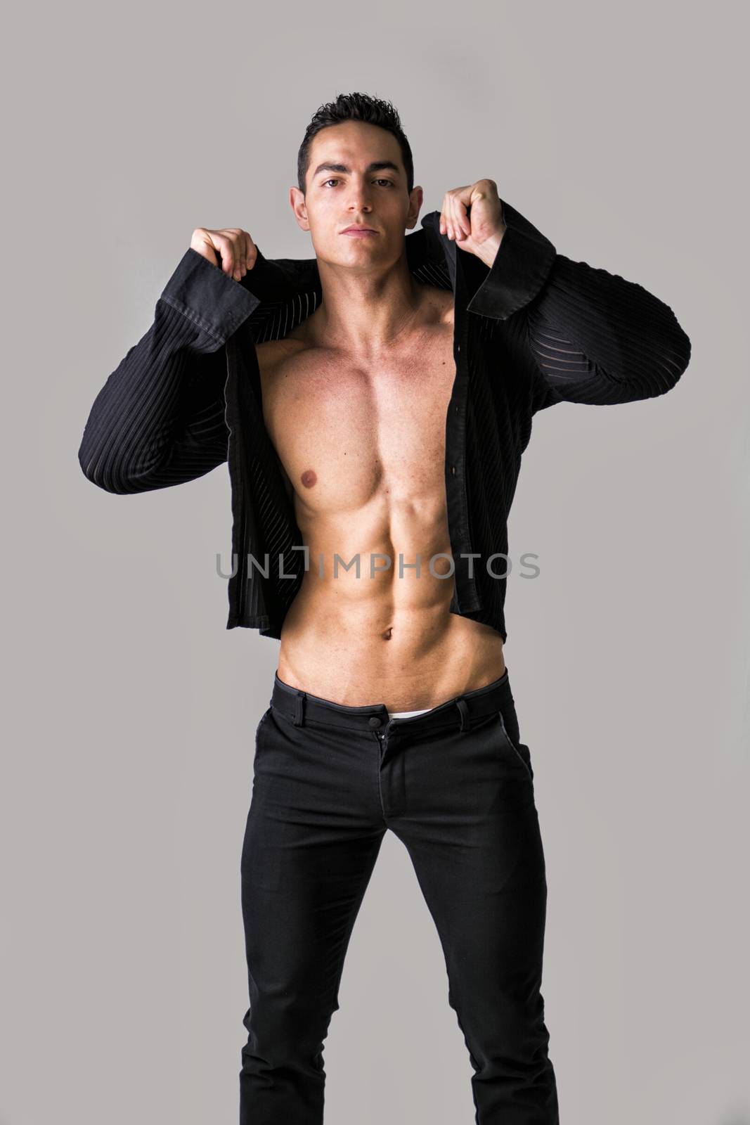Handsome muscular young man taking off shirt,on light grey background, looking at camera