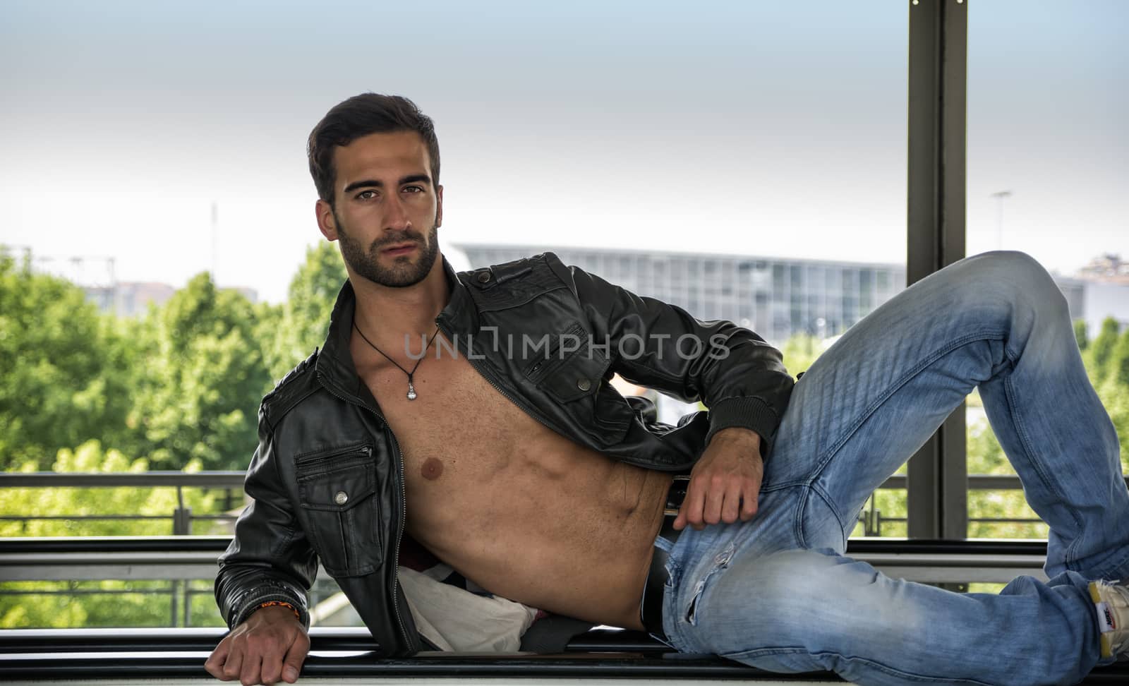 Handsome bearded young man leaning, wearing leather jacket on naked torso, outdoors in urban environment