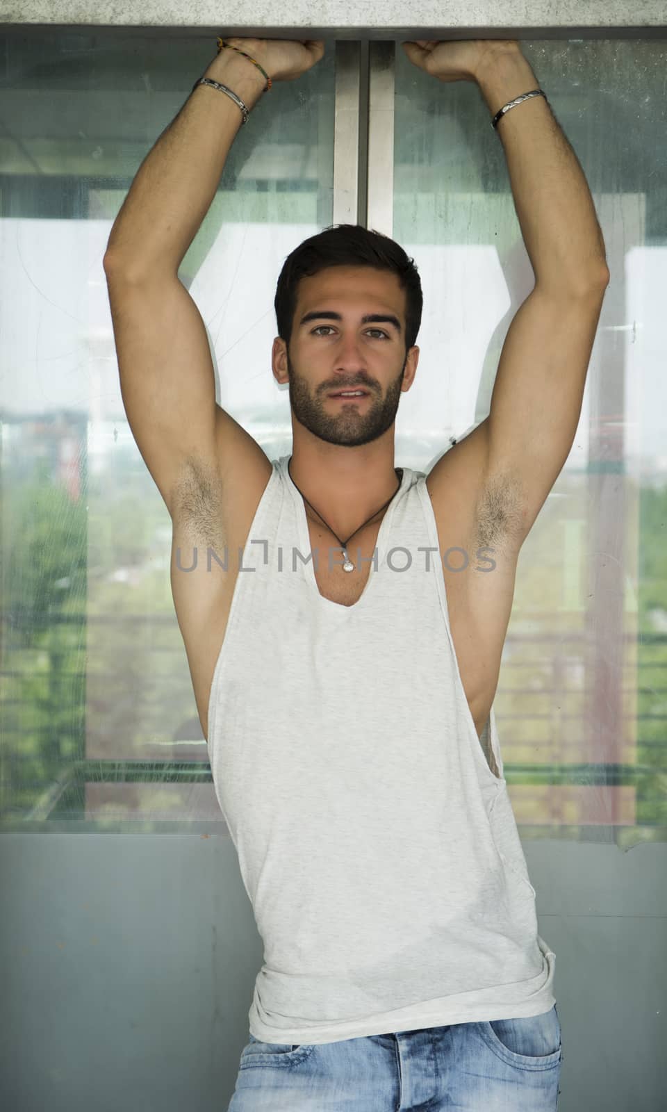 Handsome bearded young man outdoors in urban environment looking at camera, with arms up above head