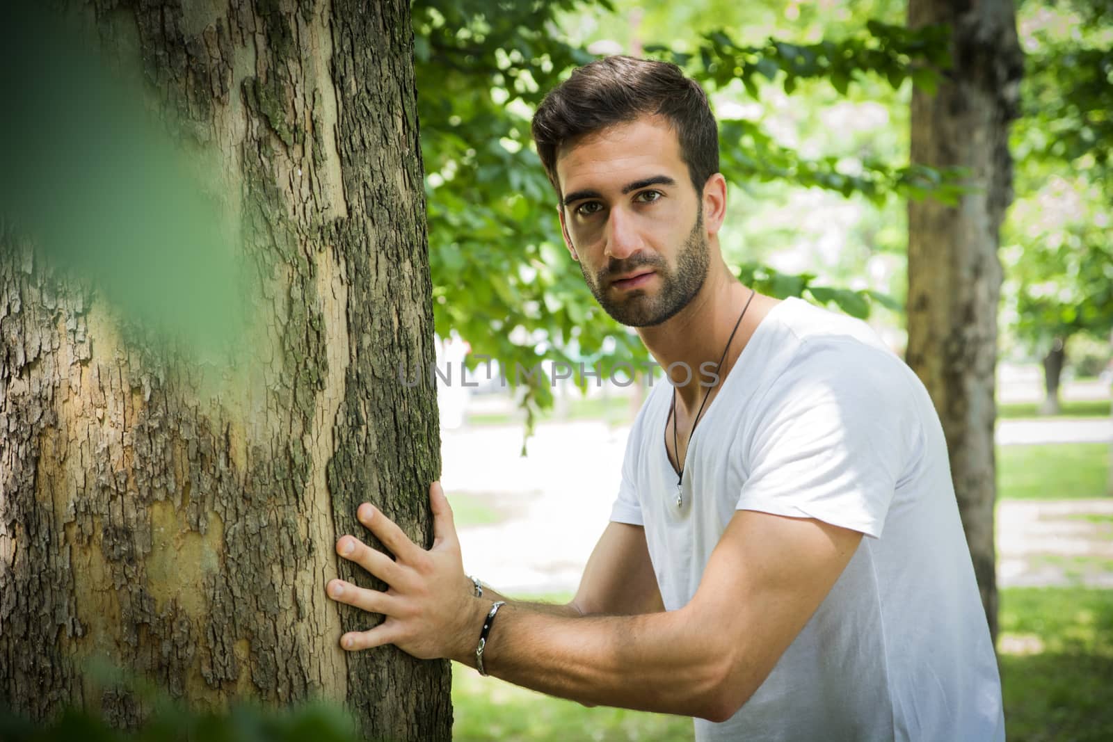 Handsome young man leaning against tree as if he were pushing it