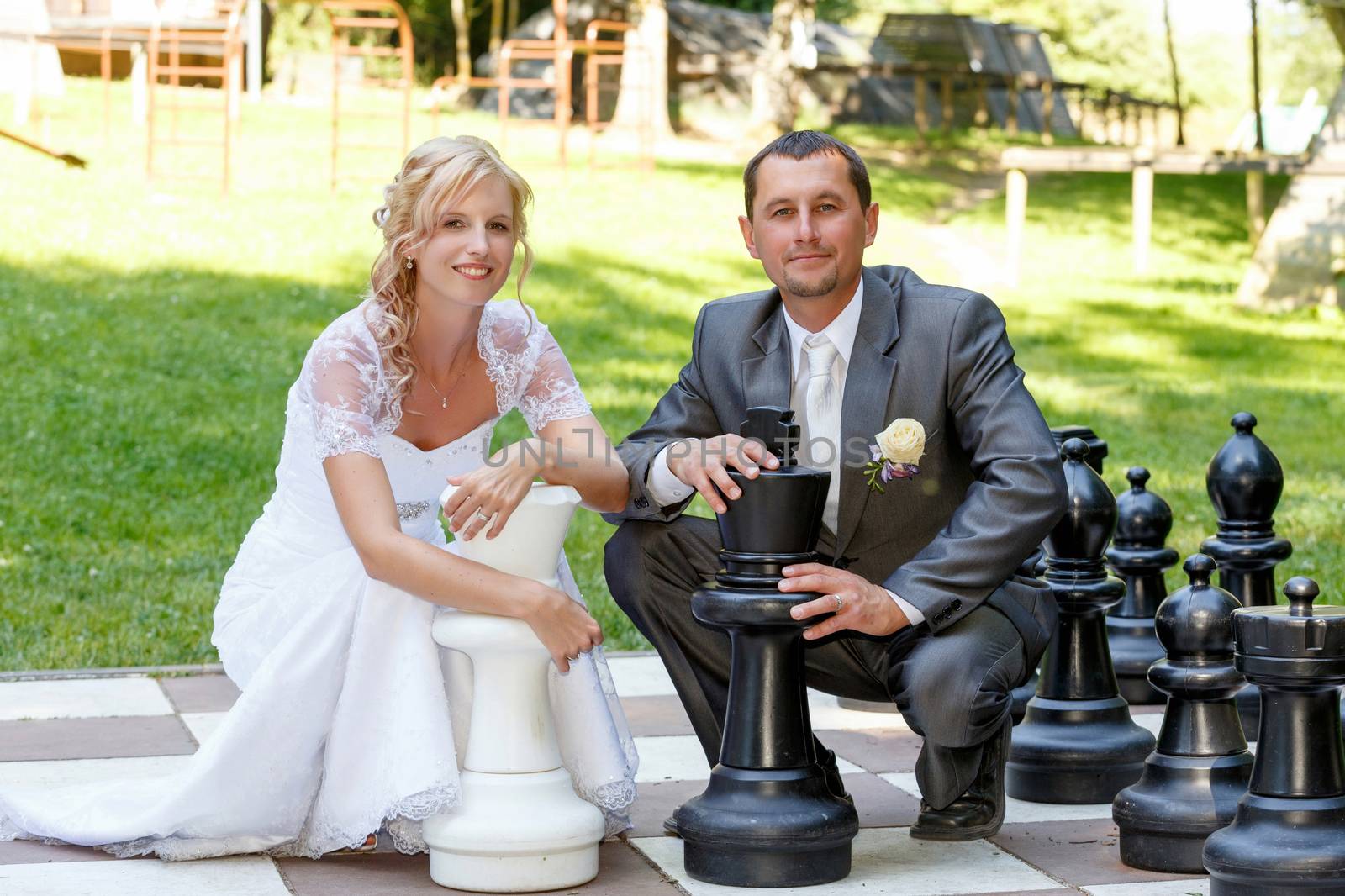 beautiful young wedding couple, blonde bride and her groom with outdoor ches, bride holding white chess checkers and groom black king