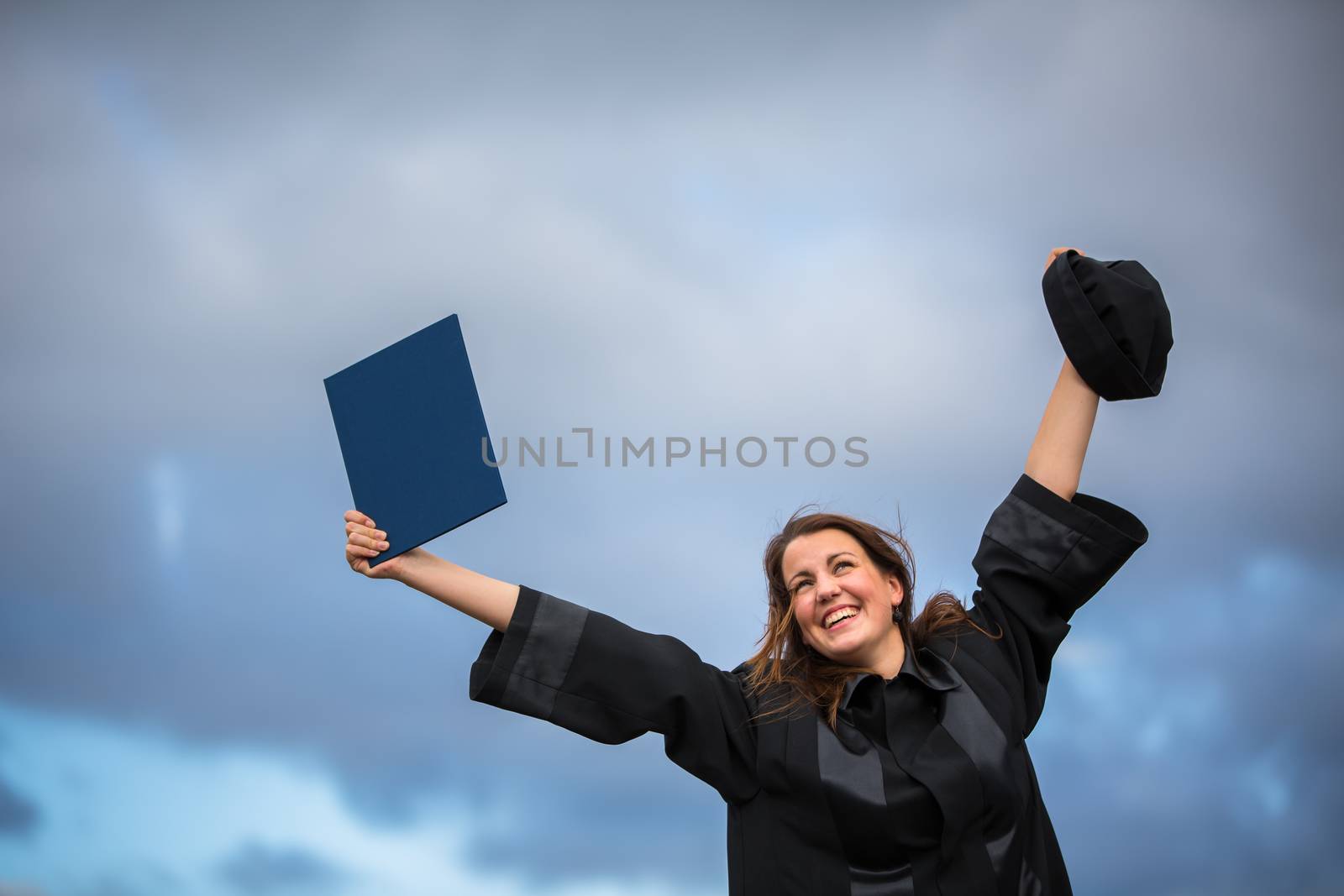 Pretty, young woman celebrating joyfully her graduation - spreading wide her arms, holding her diploma, savouring her success (color toned image; shallow DOF)