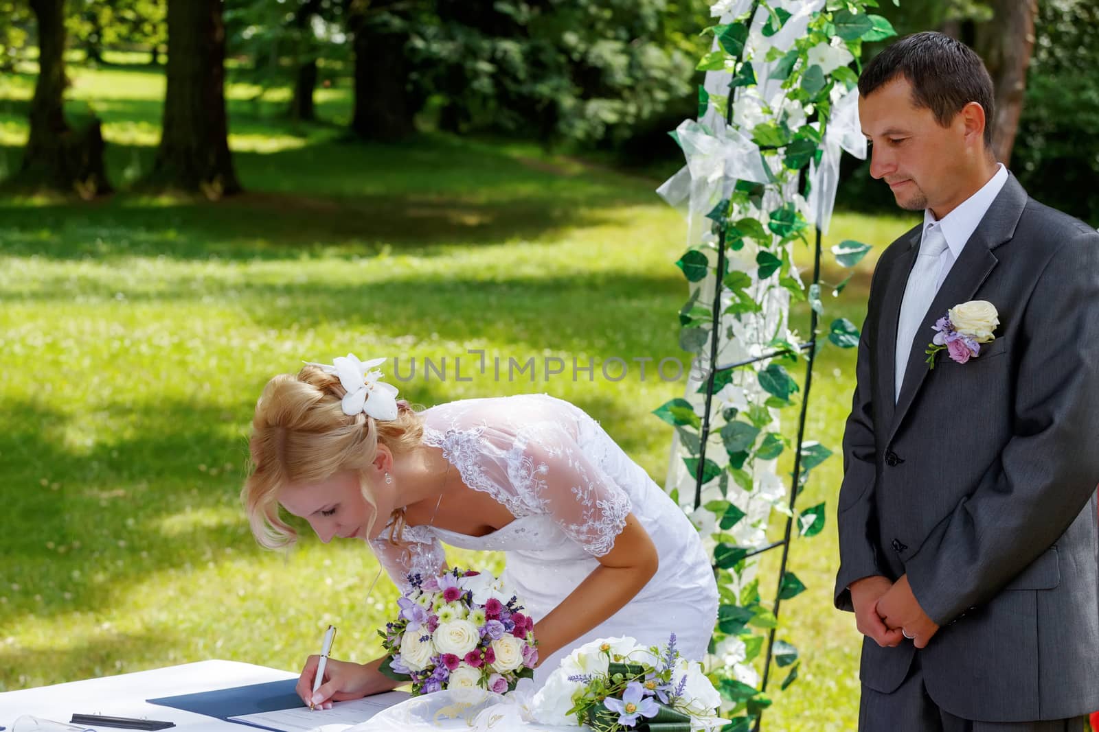 beautiful blonde smiling bride signed wedding contract against green grass outdoor, groom looking on her