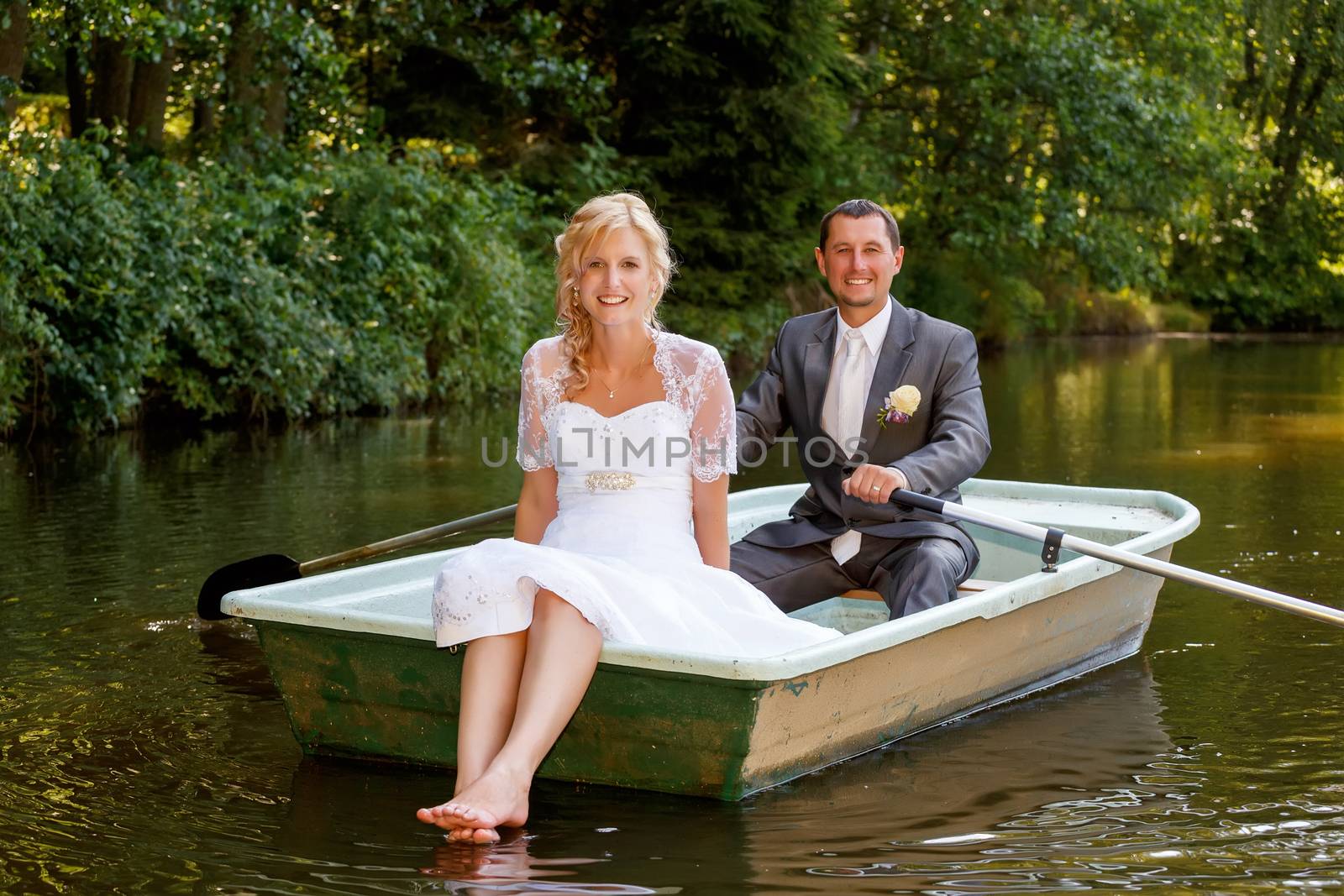 beautiful young wedding couple, blonde bride with flower and her groom just married on small boat at pond with evening sun