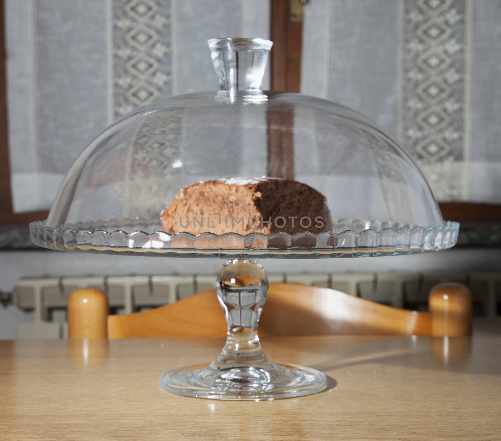 Chocolate cake inside a glass stand, over kitchen table