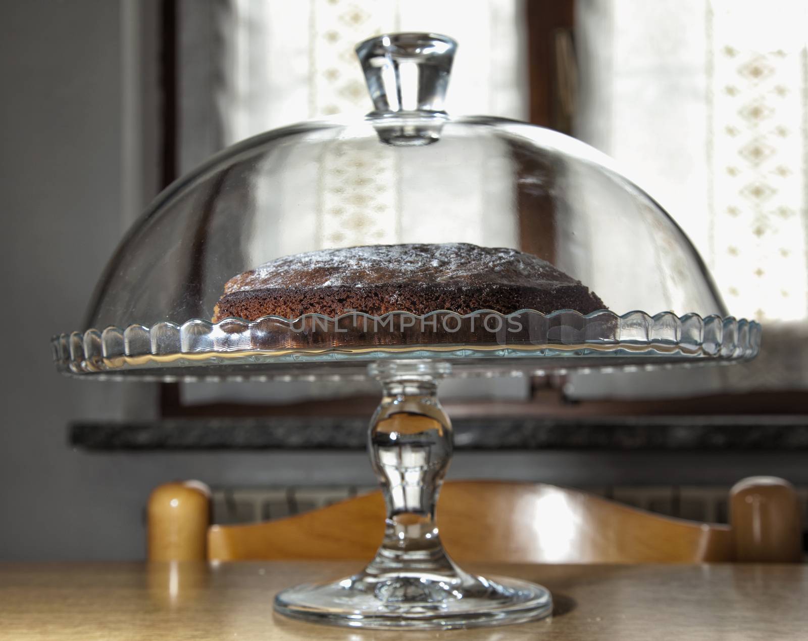 Chocolate cake inside a glass stand, over kitchen table