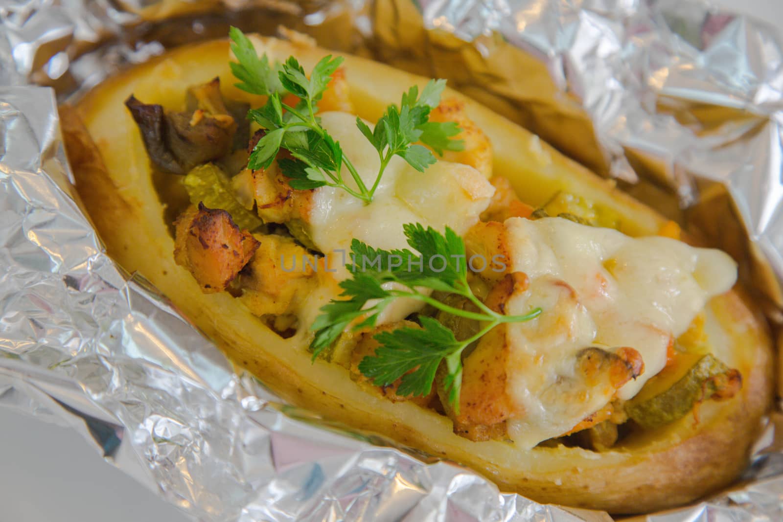 Baked jacket potato with vegetables,chicken filet,fresh parsley and cheese in the aluminium foil  on the white plate