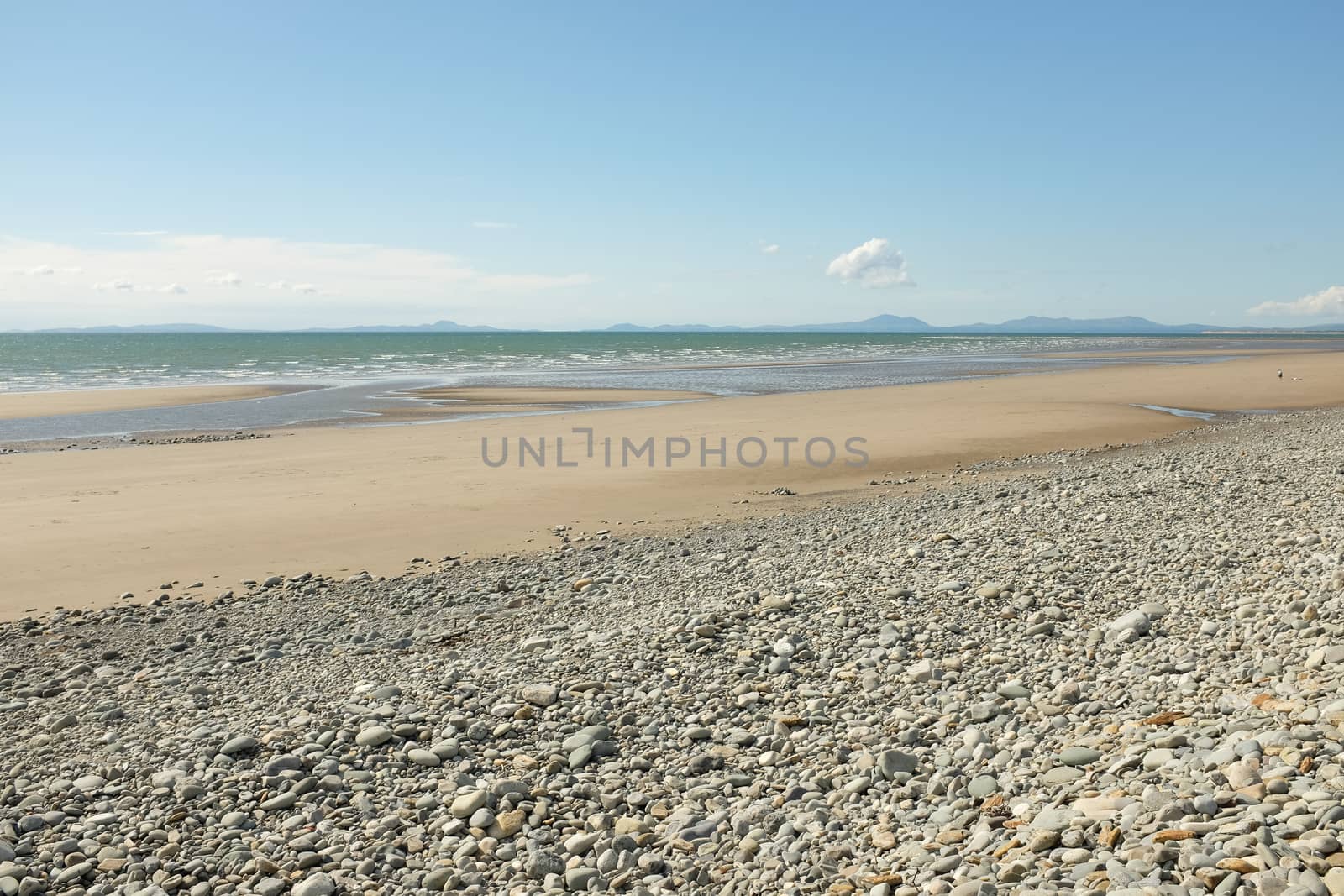 Pebbles lead down to a deserted sandy beach and sea with mountains in the distance.