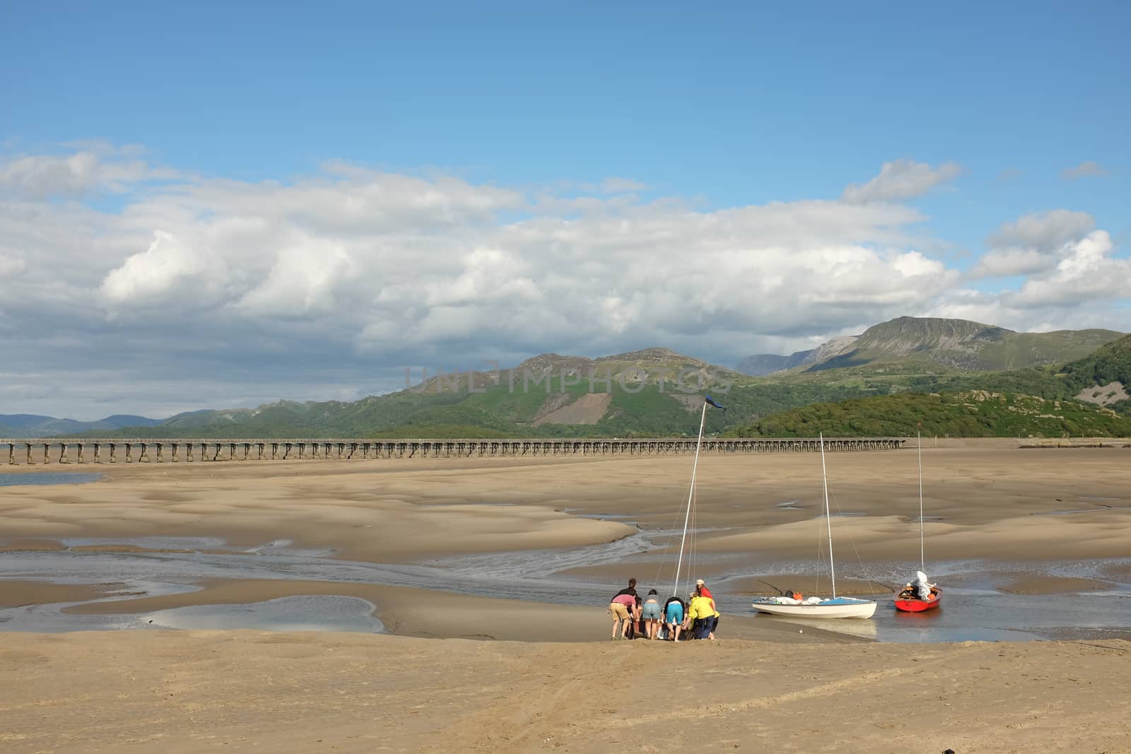 People preparing sail boats on the sand of an estuary at low tide with mountains in the distance.