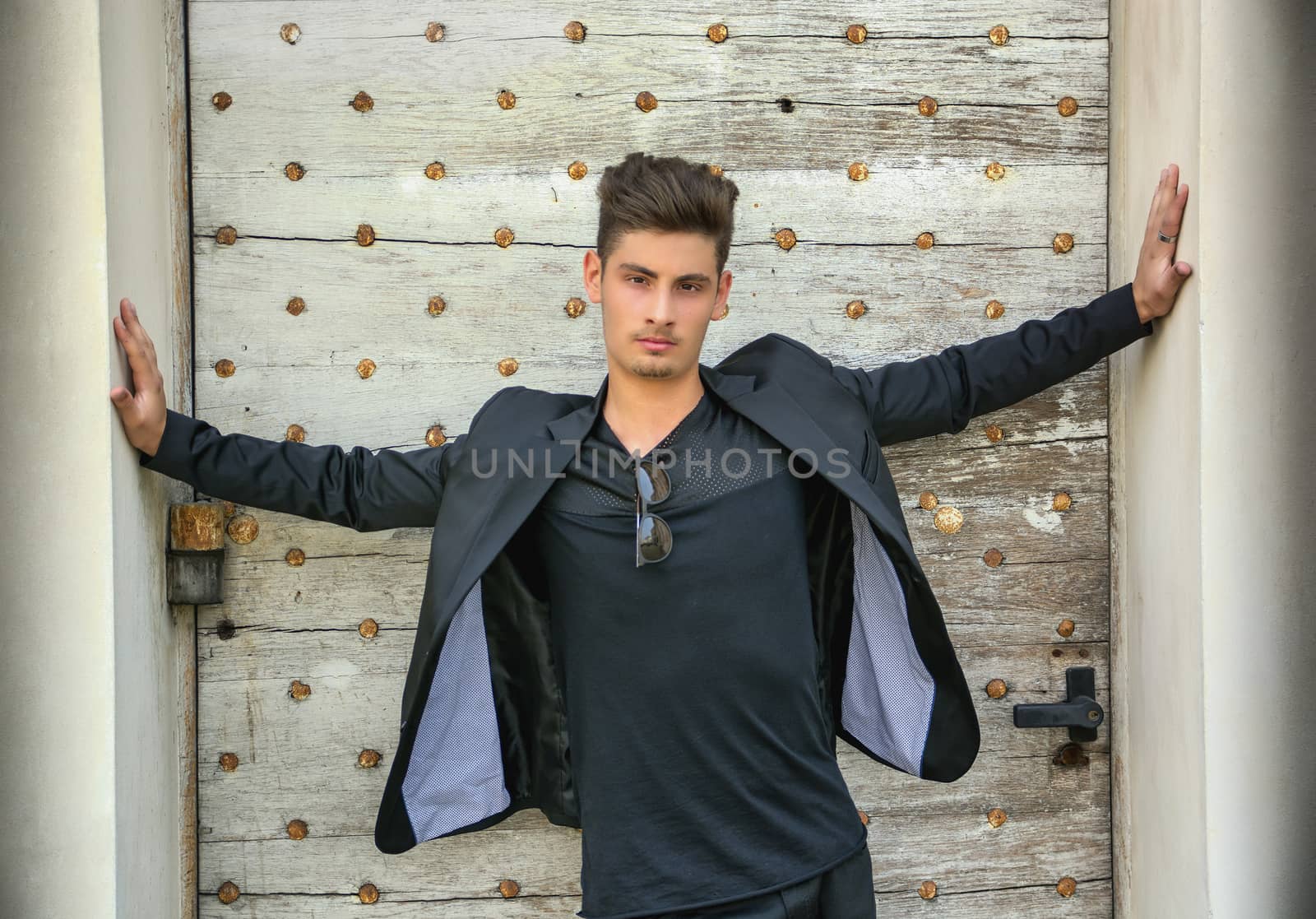 Handsome young man with arms spread open in front of old door, wearing elegant jacket, looking at camera