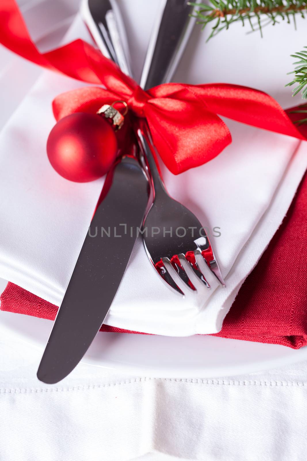 Romantic red Christmas table setting with white plates, red and white linen and silverware tied with a red ribbon and bow decorated with red Xmas baubles and evergreen natural pine foliage