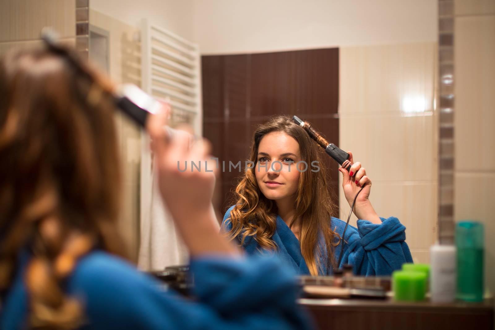 Pretty, young woman curling her hair in front of her bathroom mirror