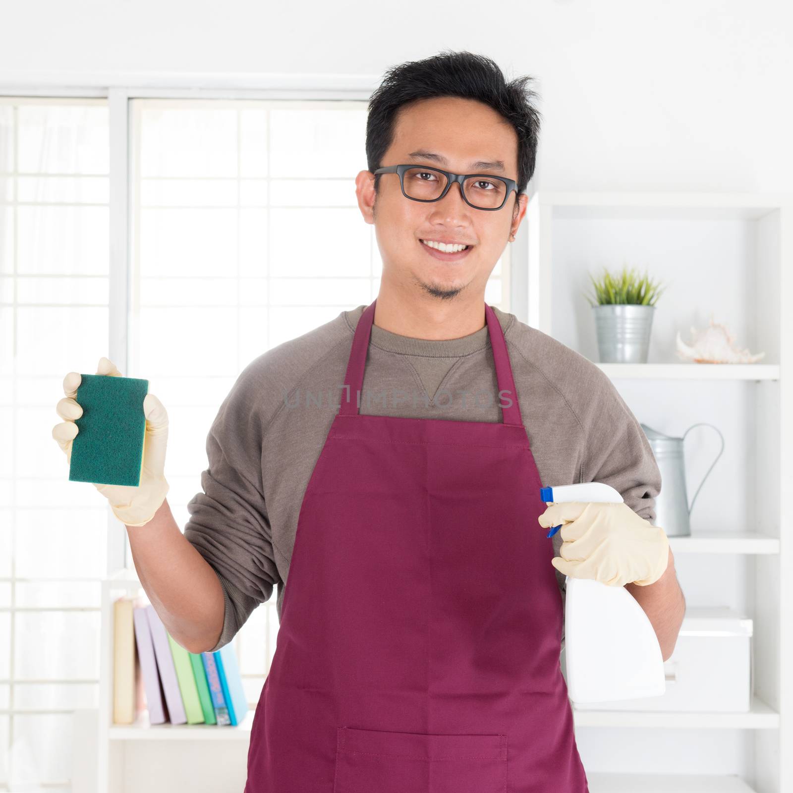 Asian man holding sponge and spray. House husband doing house chores, with interiors.