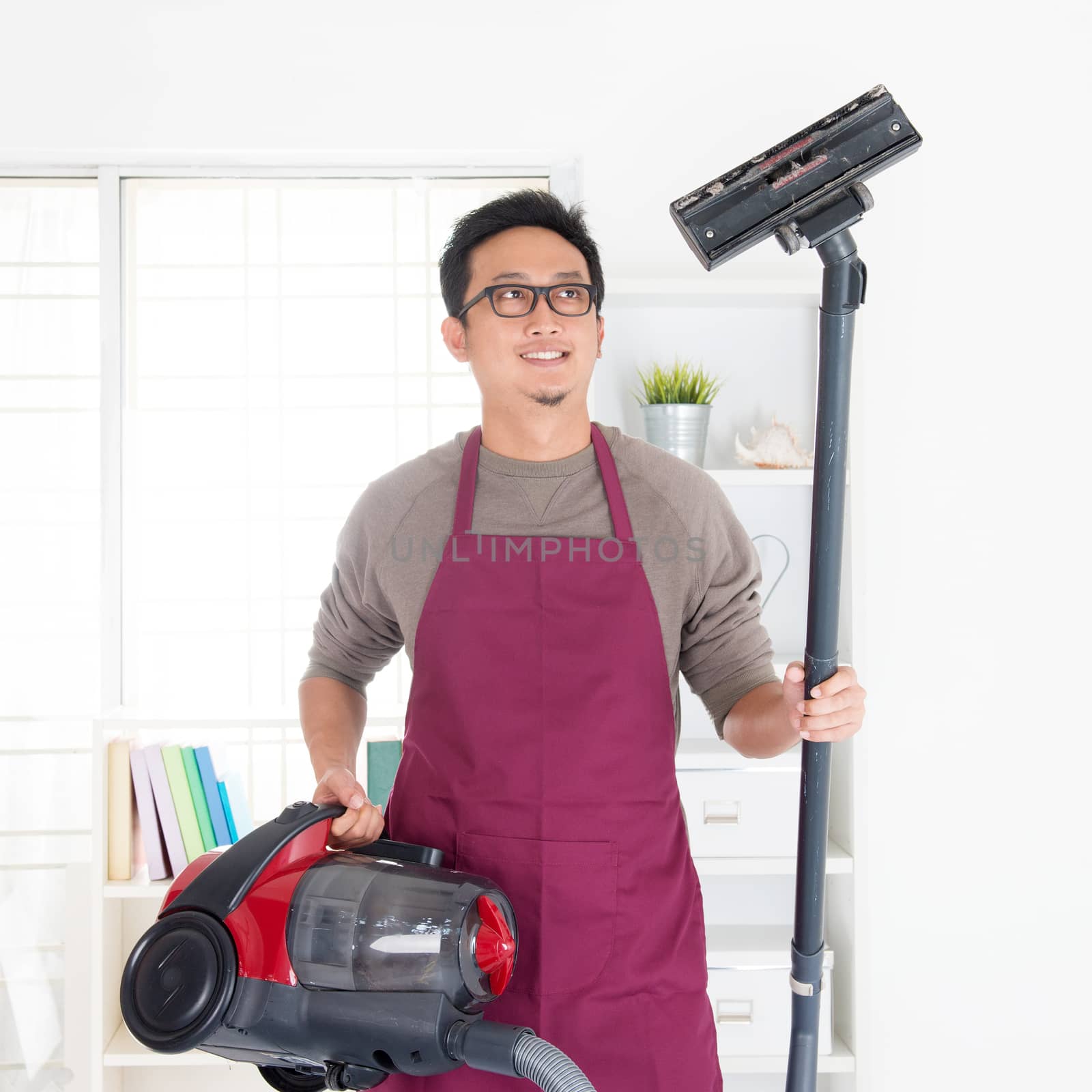 Asian man holding vacuum. House husband doing house chores, house interior.