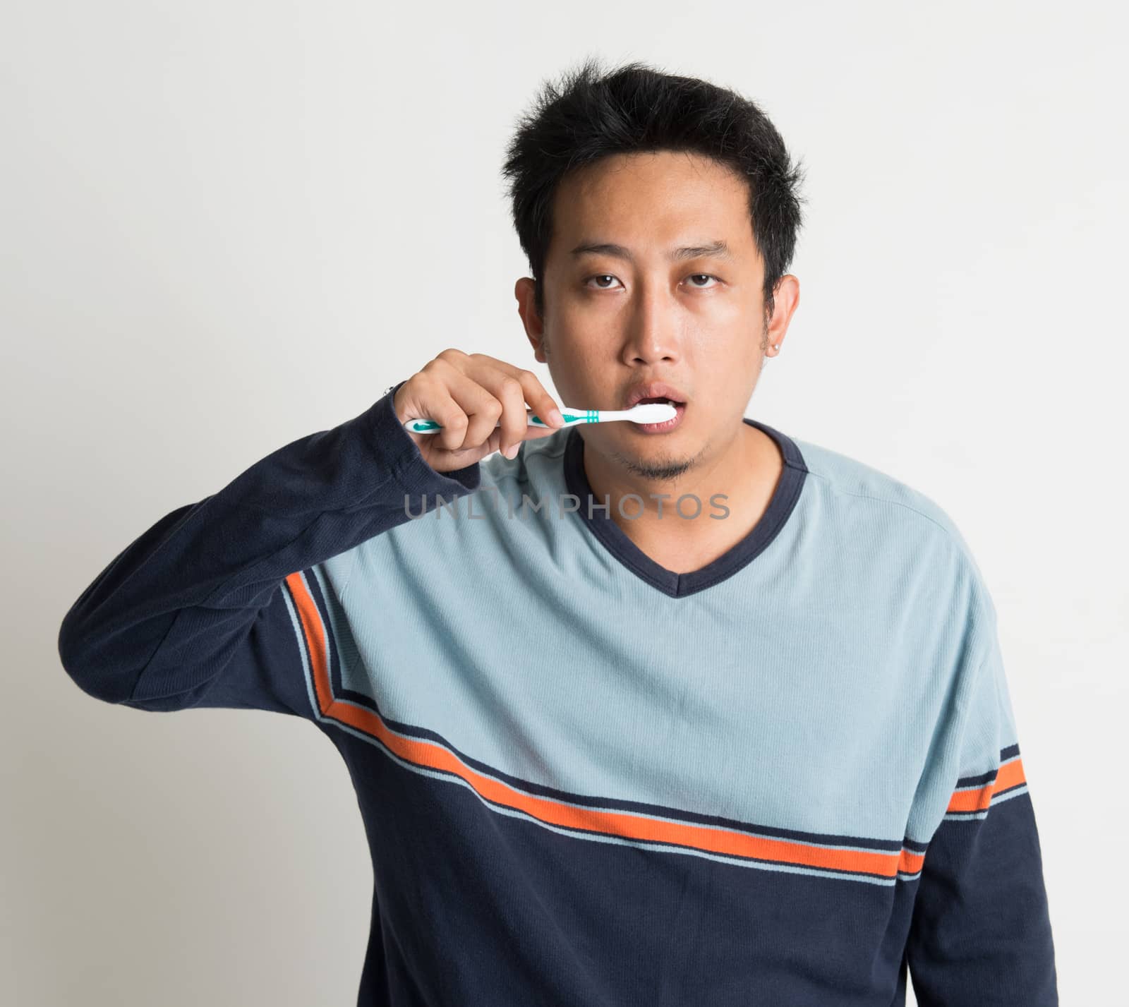 Southeast Asian male brushing teeth with sleepy eyes in a morning, on plain background