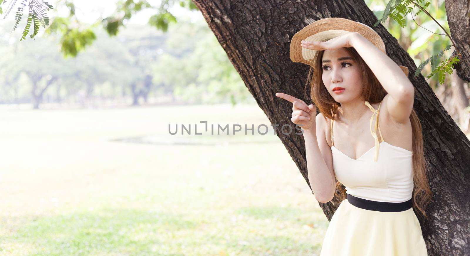 Portrait Asian woman under a tree. Woman with long hair wearing a hat is pointing forward.