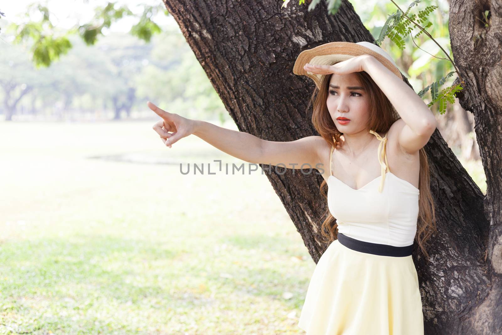 Portrait Asian woman under a tree. Woman with long hair wearing a hat is pointing forward.