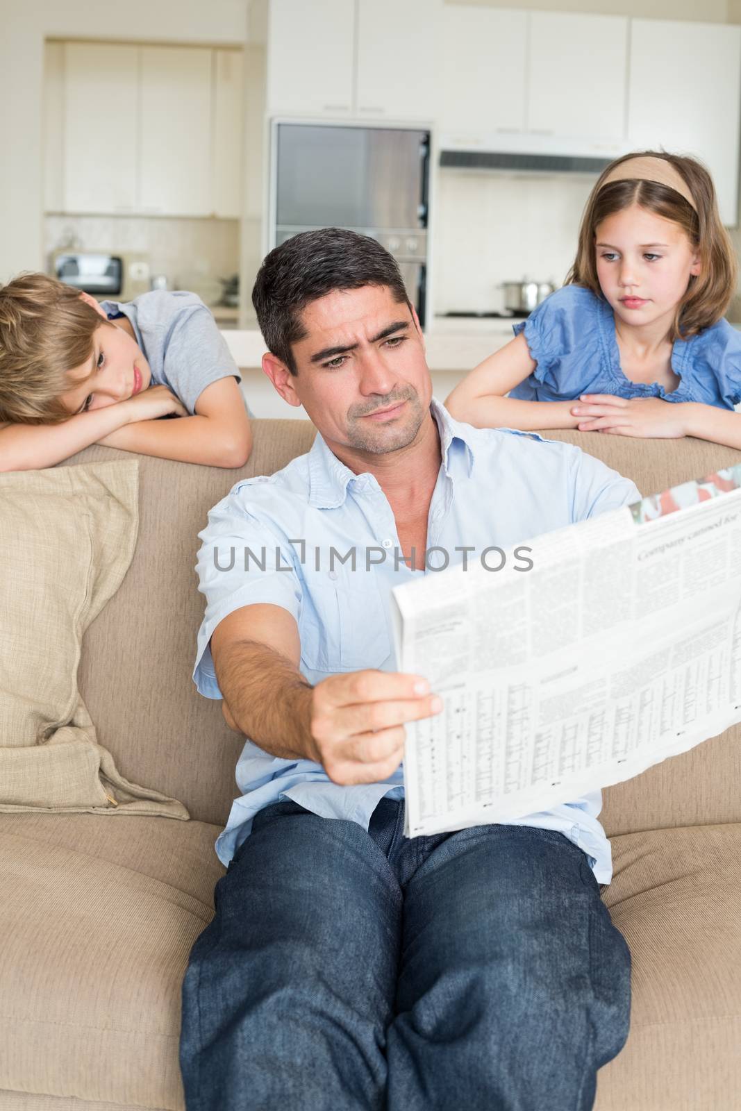 Bored children looking at father reading newspaper in house