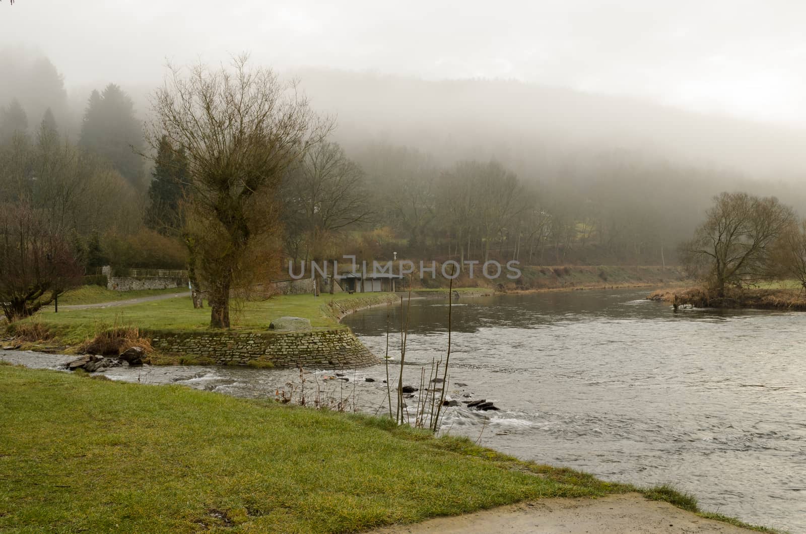 Low hanging clouds produce a fog effect in the mountains behind this river in Belgium