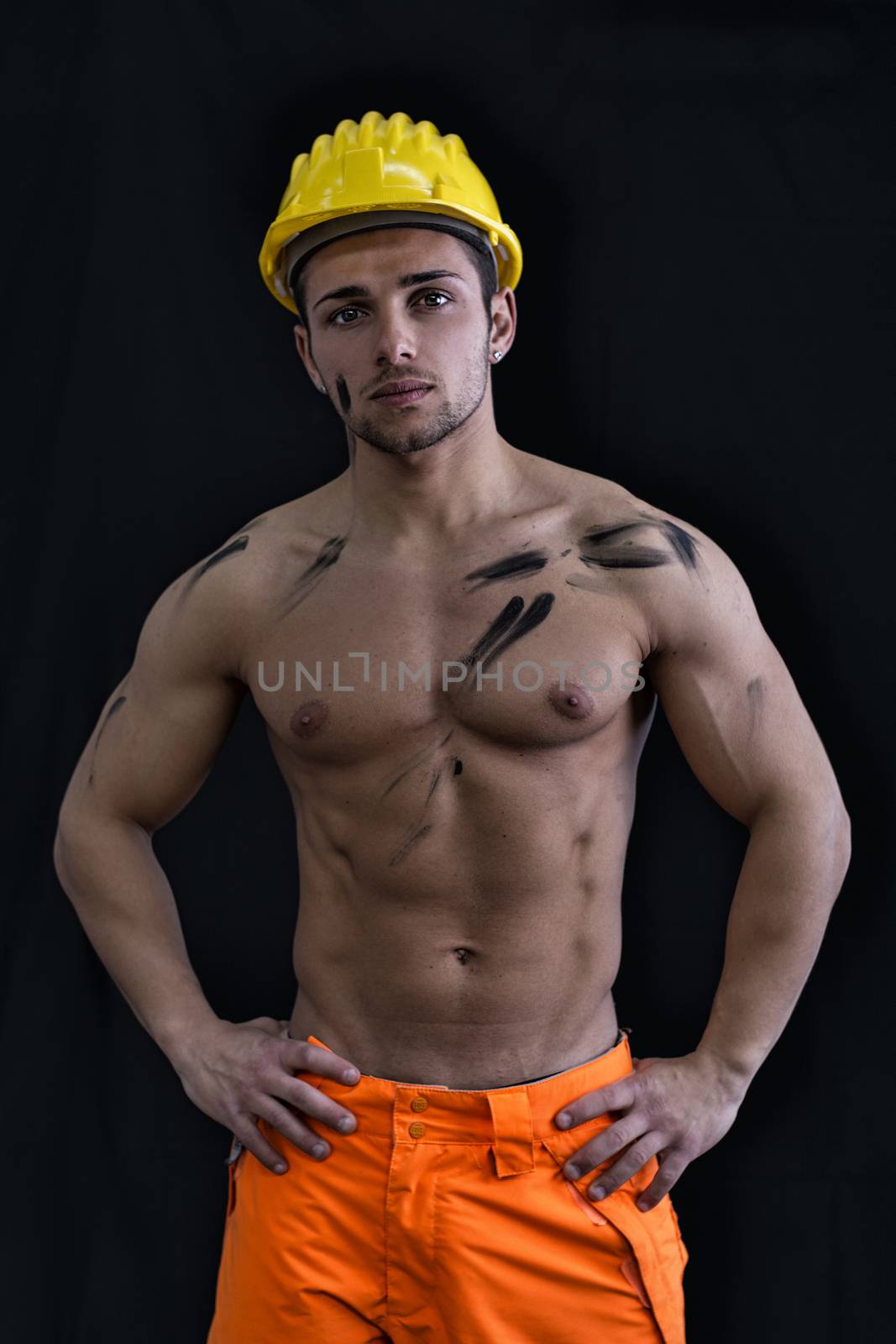 Muscular young construction worker shirtless wearing hardhat, isolated on black background