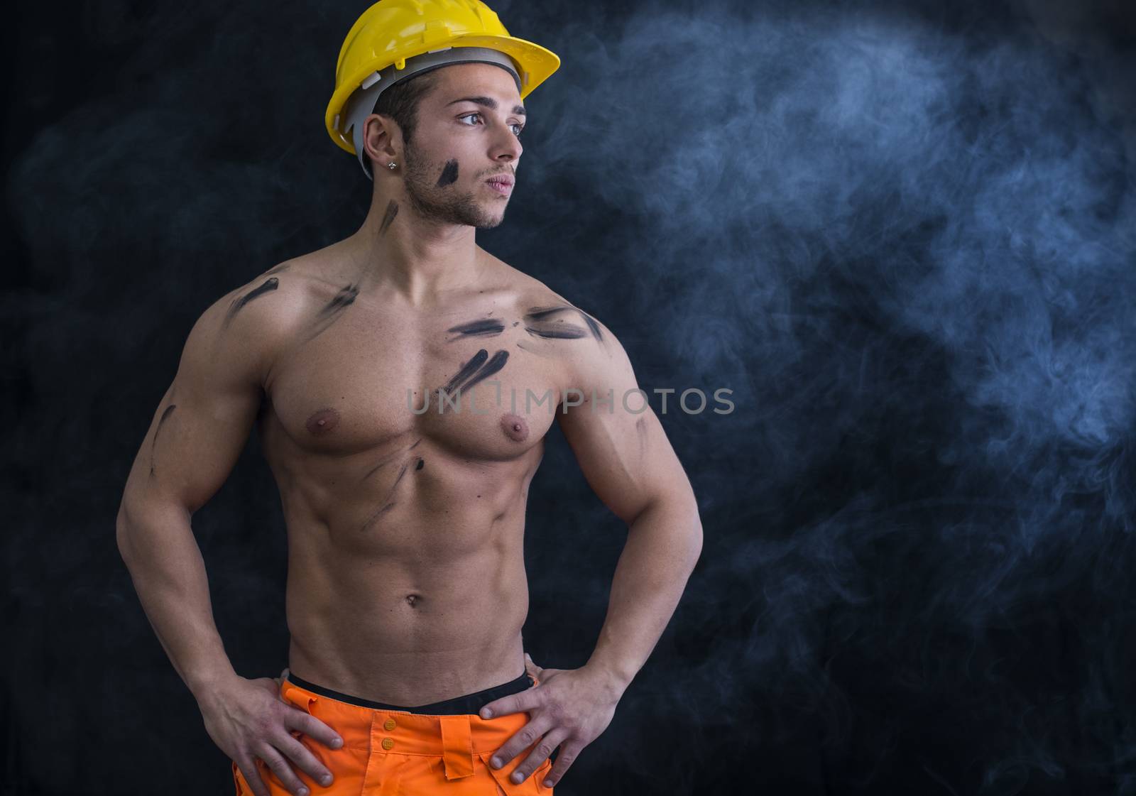 Muscular young construction worker shirtless wearing hardhat, dark background with smoke