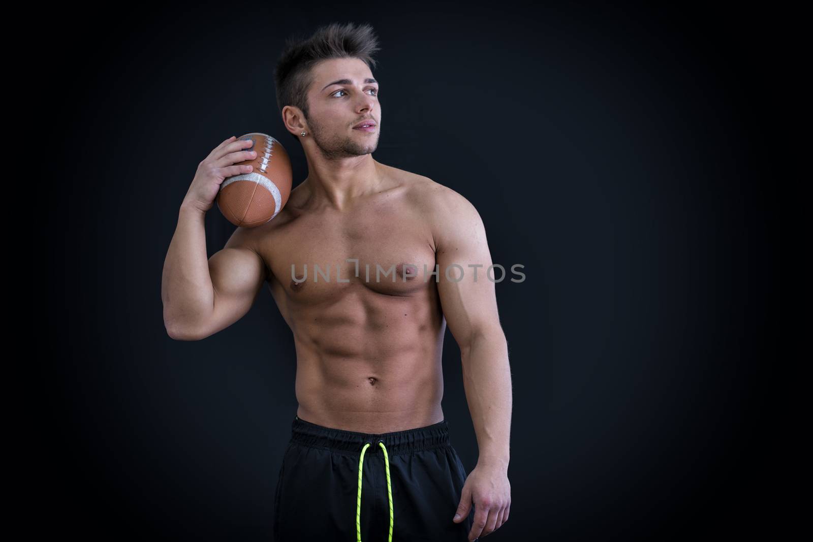 Muscular american football player standing with ball in hand, on dark background