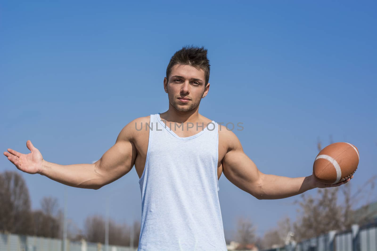 Muscular american football player standing with ball in hand, arms open. Outdoors shot