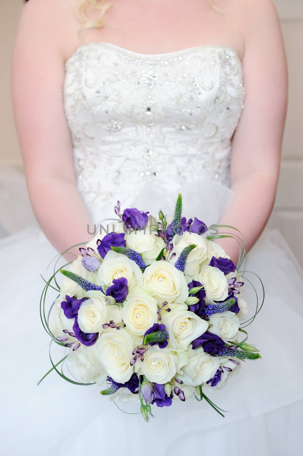 Bride holding bouquet of white and purple flowers on wedding day showing dress detail closeup