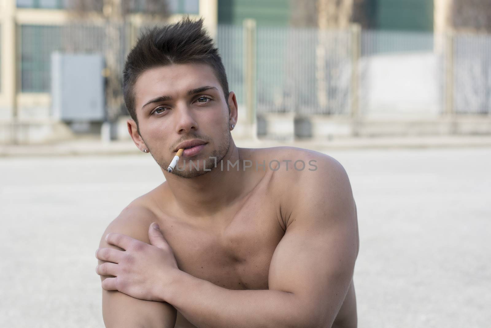 Shirtless muscular young man smoking cigarette outside looking at camera