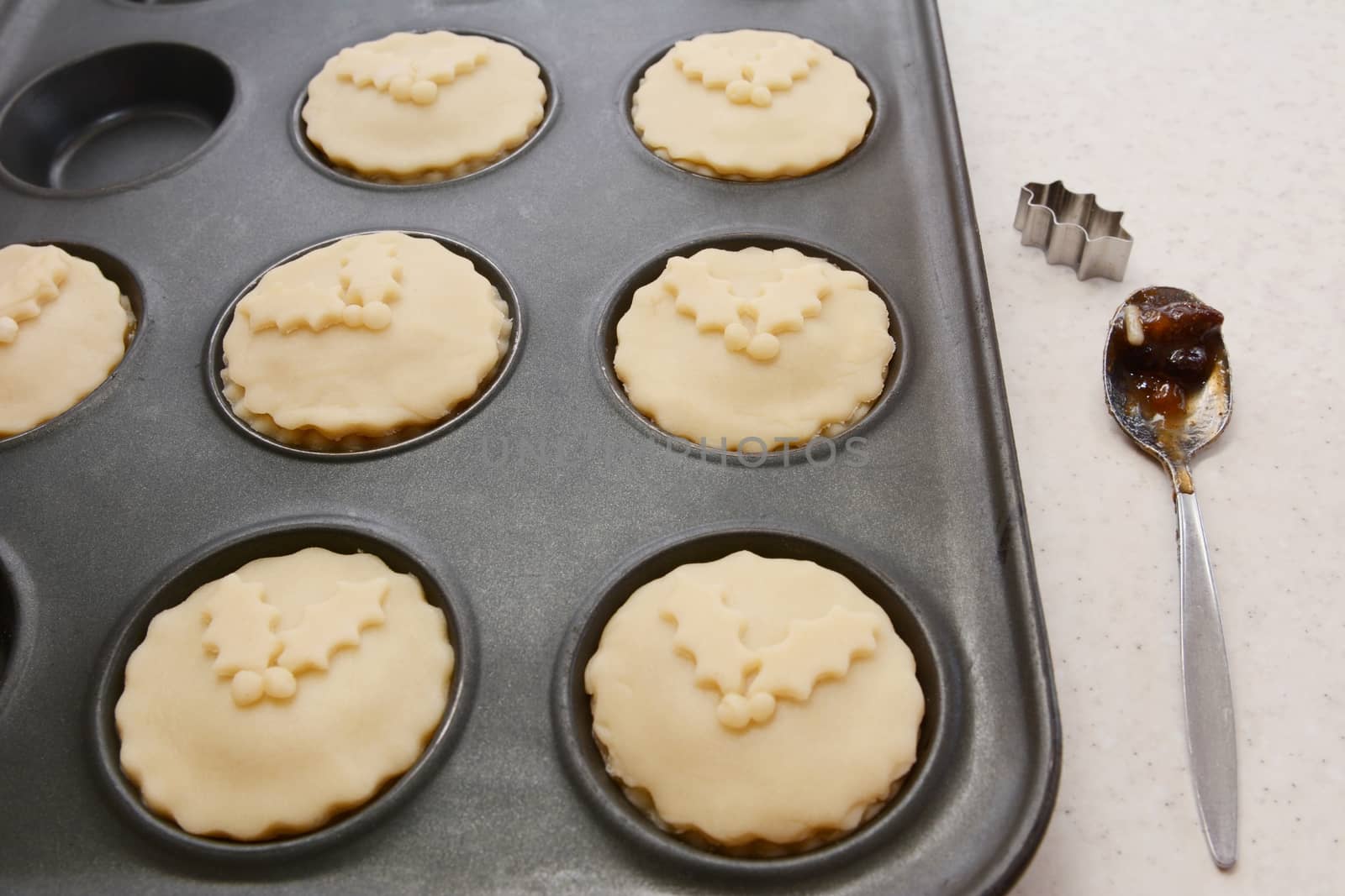 Mince pies ready to be baked, with teaspoon of mincemeat and a holly leaf cutter on the worktop