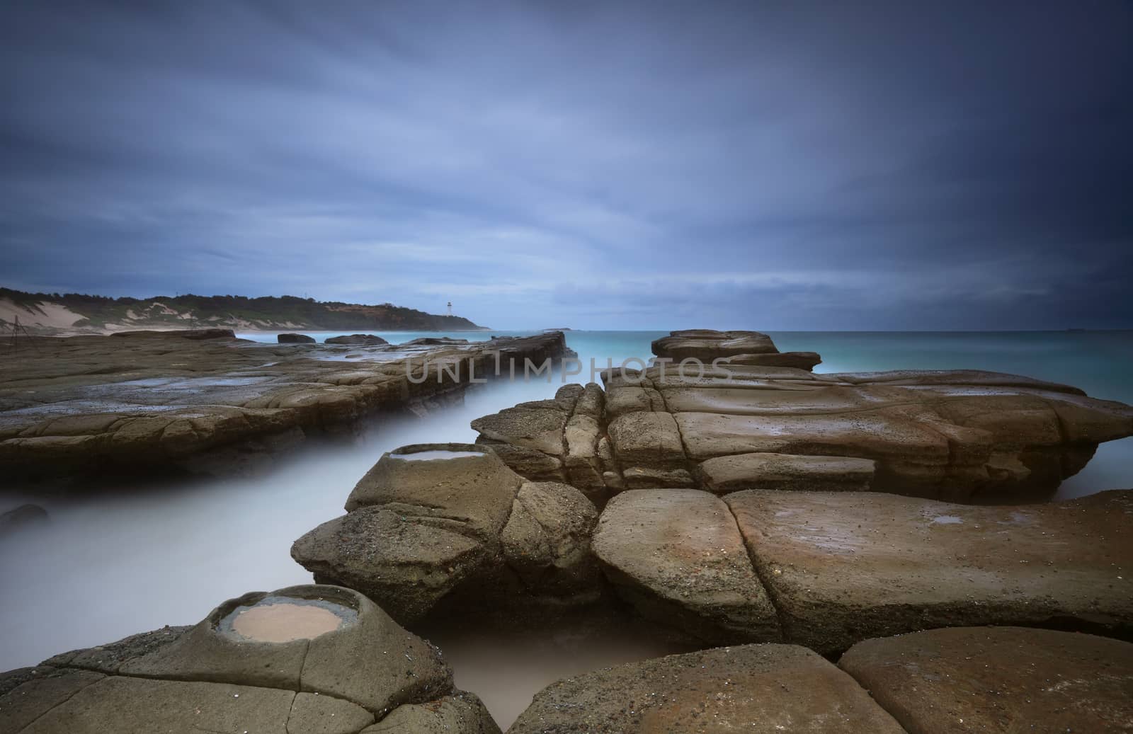 Weathered rocks, ocean channel and rock craters at Soldiers Beach Australia under a stormy dark sky.  Norah Head lighthouse in distance.