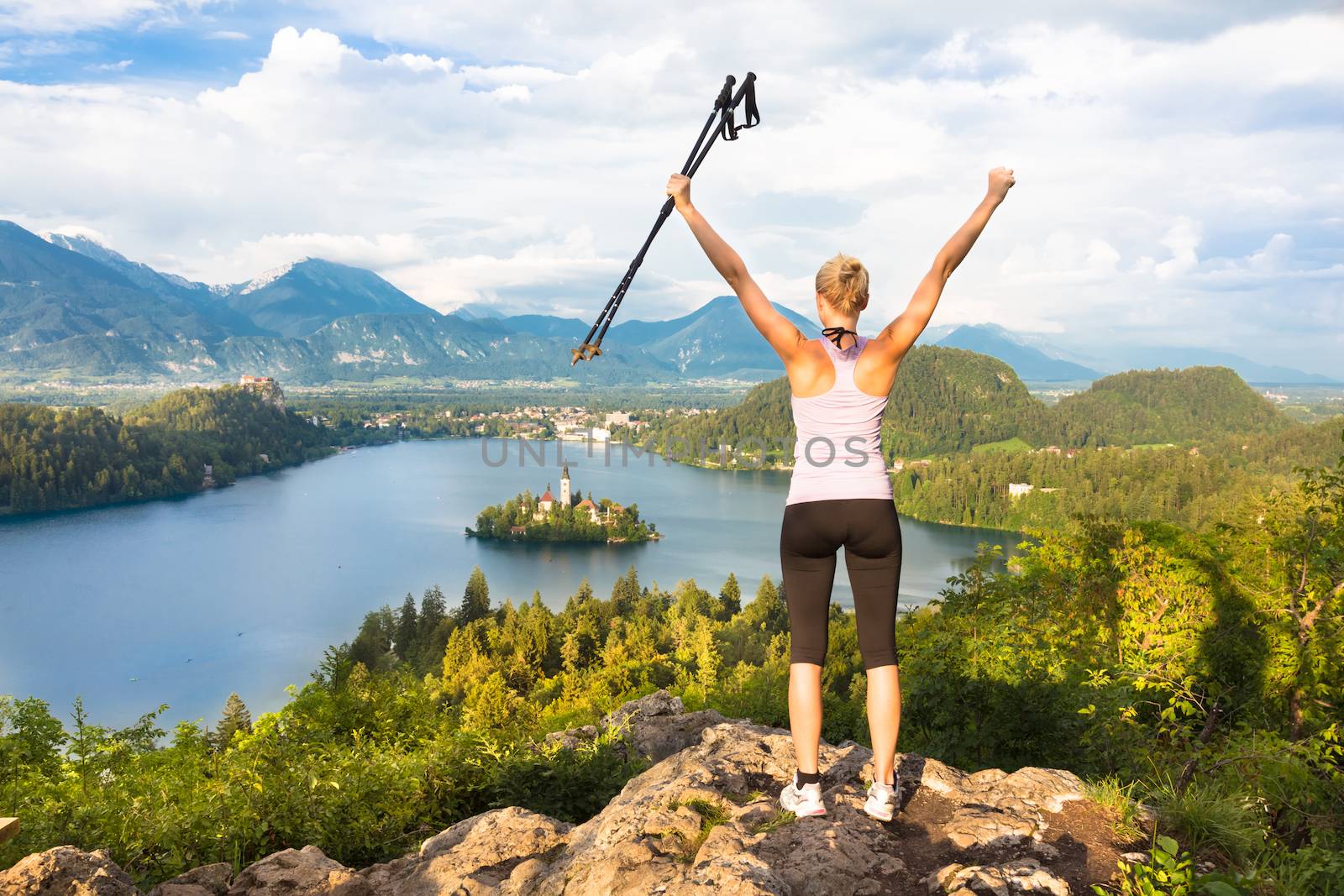 Made it! Young spoty active lady with hiking sticks and hands rised admiring beautiful nature around Bled Lake in Julian Alps, Slovenia. 