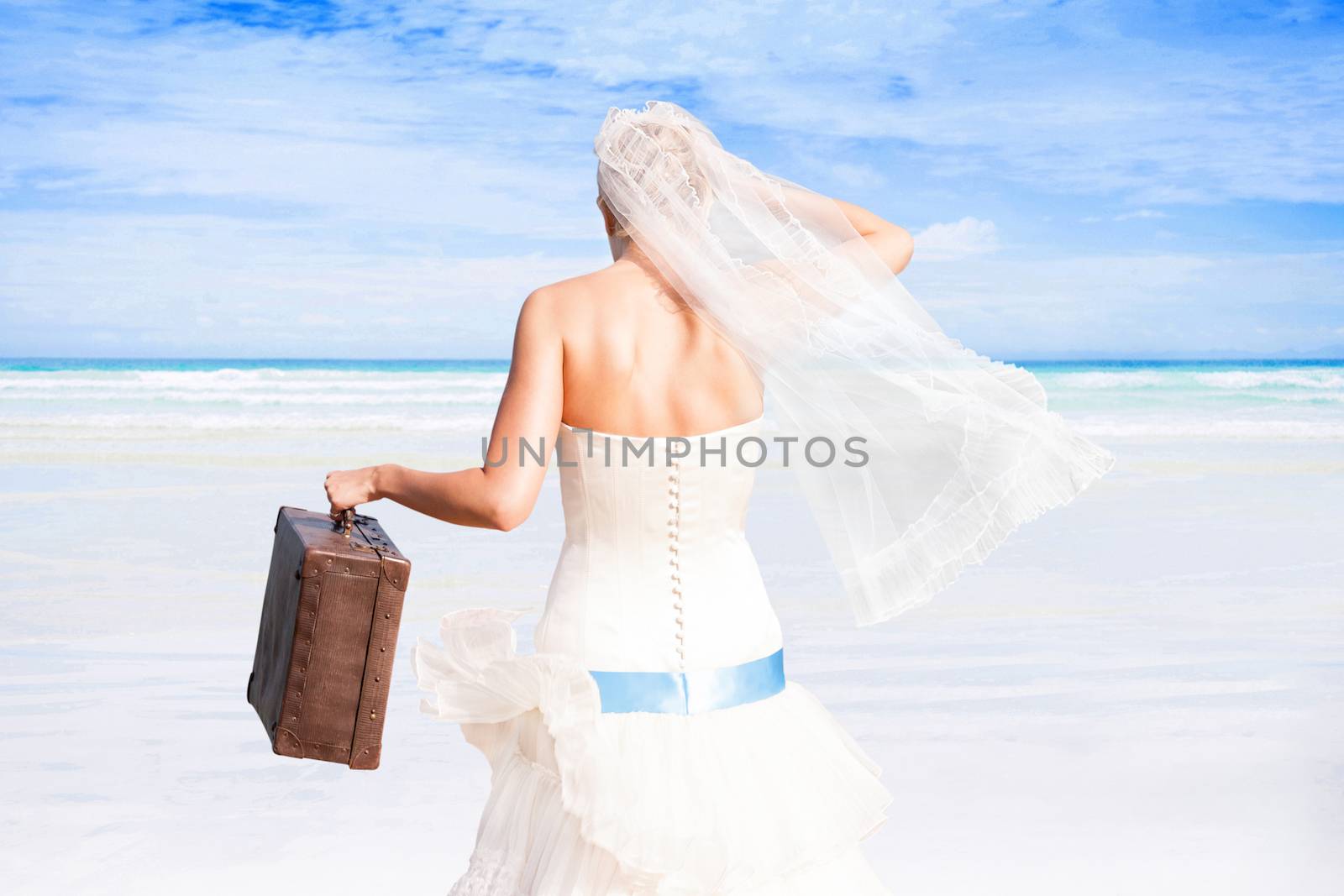 Beautiful bride with antique suitcase in white wedding dress on white sand beach with turquoise ocean on sunny day. Luxurious honeymoon, romantic wedding.