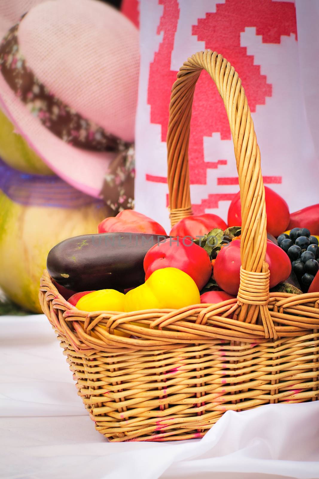 On a white towel there is a beautiful basket with tomatoes, eggplants, pepper, grapes for sale at fair.