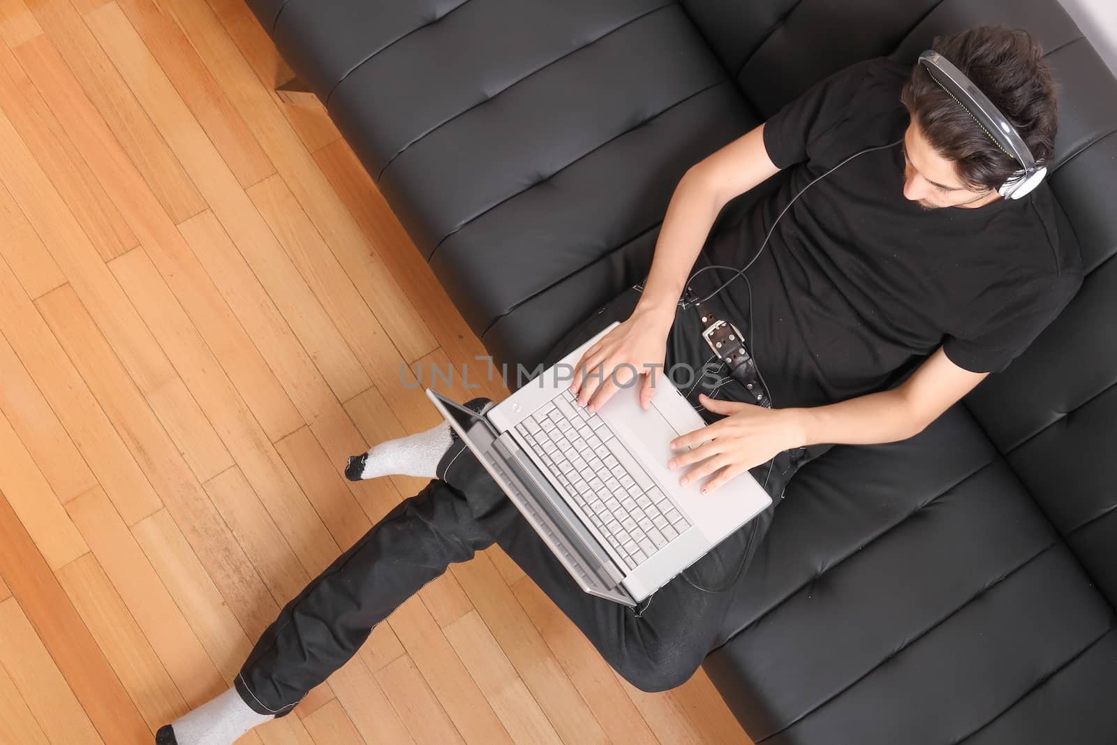 A young hispanic man with a laptop on the Sofa listening Music with headphones.