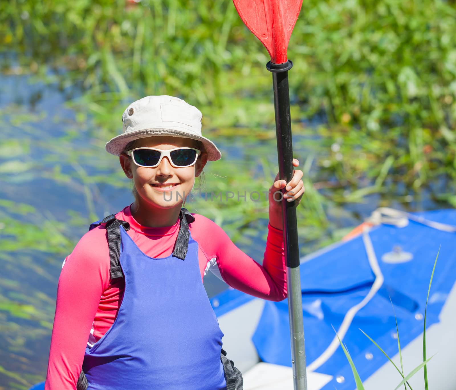 Portrait of happy cute girl holding paddle near a kayak on the river, enjoying a lovely summer day